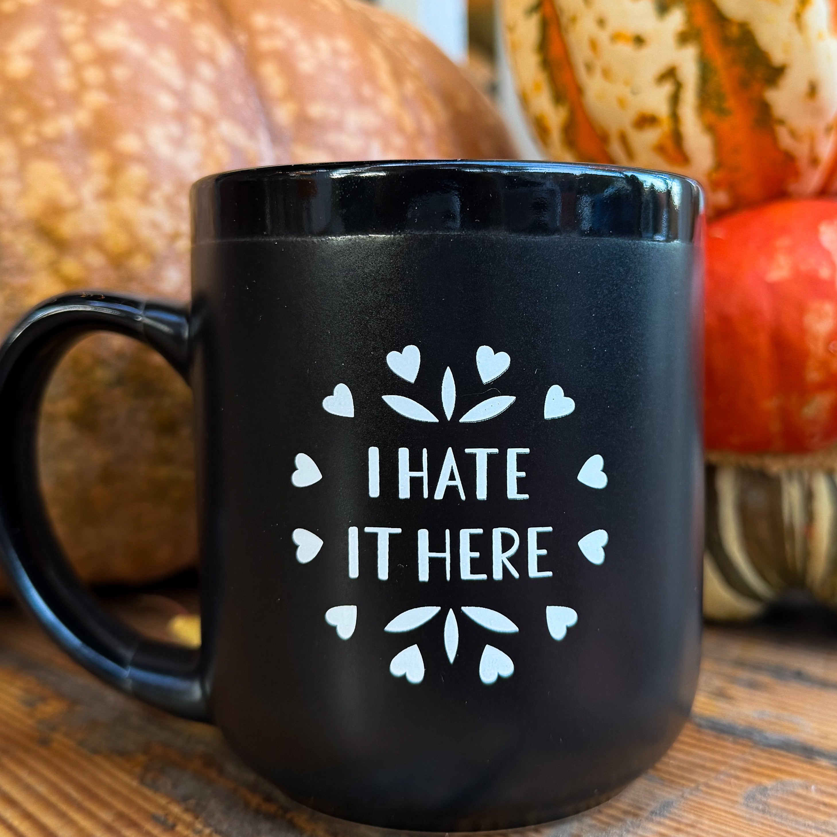 A black ceramic mug that says 'I HATE IT HERE' in white surrounded by a circle of hearts and some leafy embellishments with pumpkins in the background.