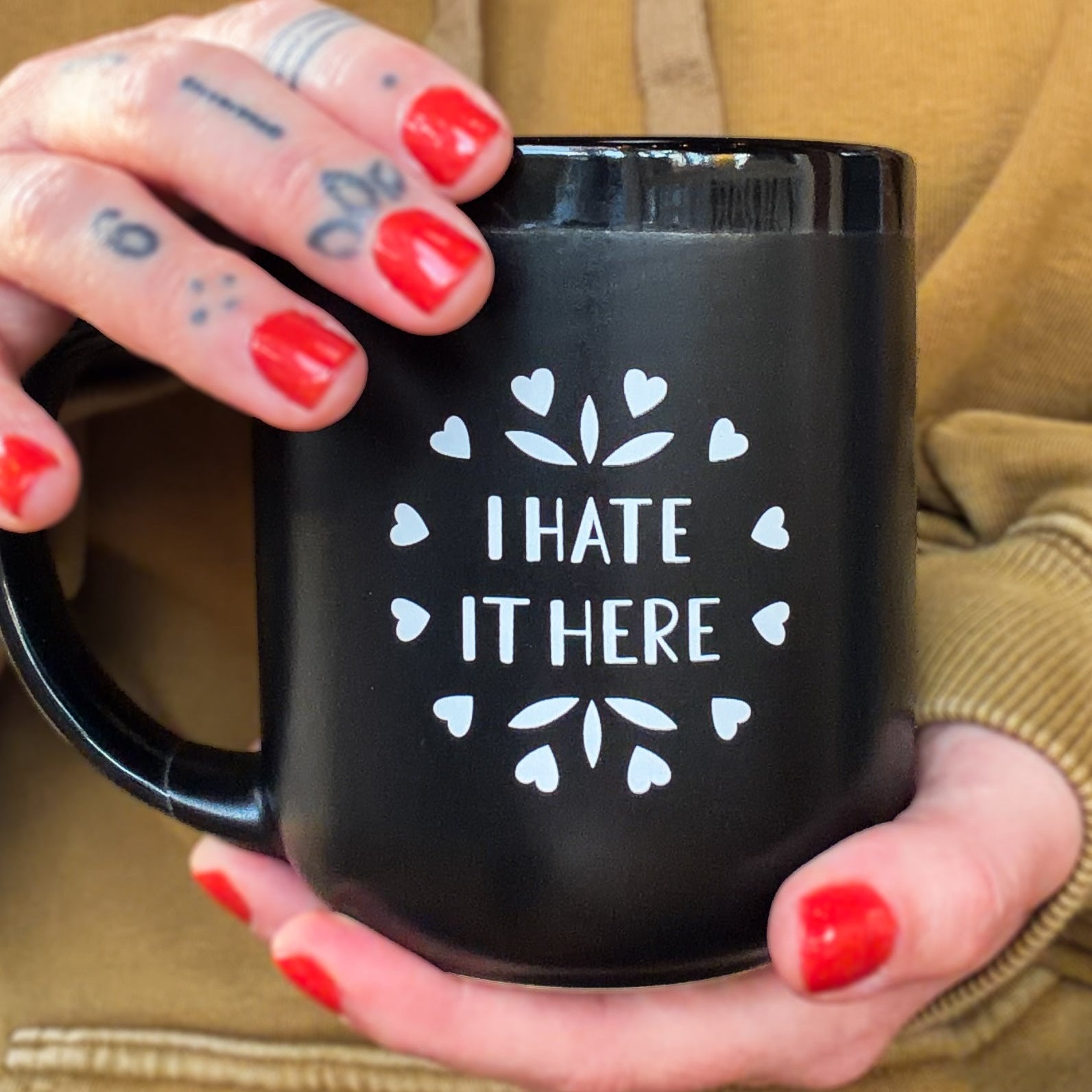 A black ceramic mug that says 'I HATE IT HERE' in white surrounded by a circle of hearts and some leafy embellishments being held by a hand with tattoos and red nails.