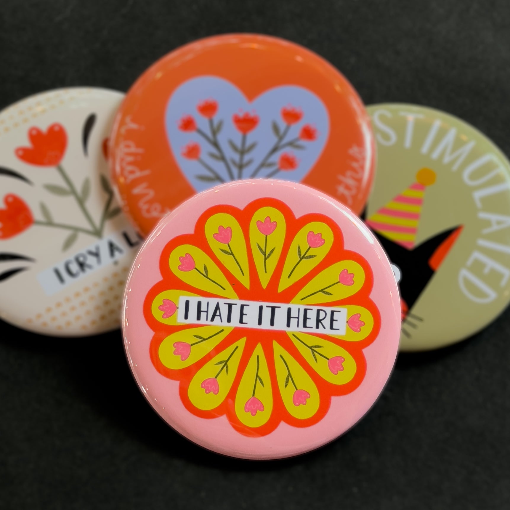 A round pinback button featuring a pink background with a circle of pink flowers on a yellow and red background and the text 'I HATE IT HERE'. The pin is surrounded by other pin designs.