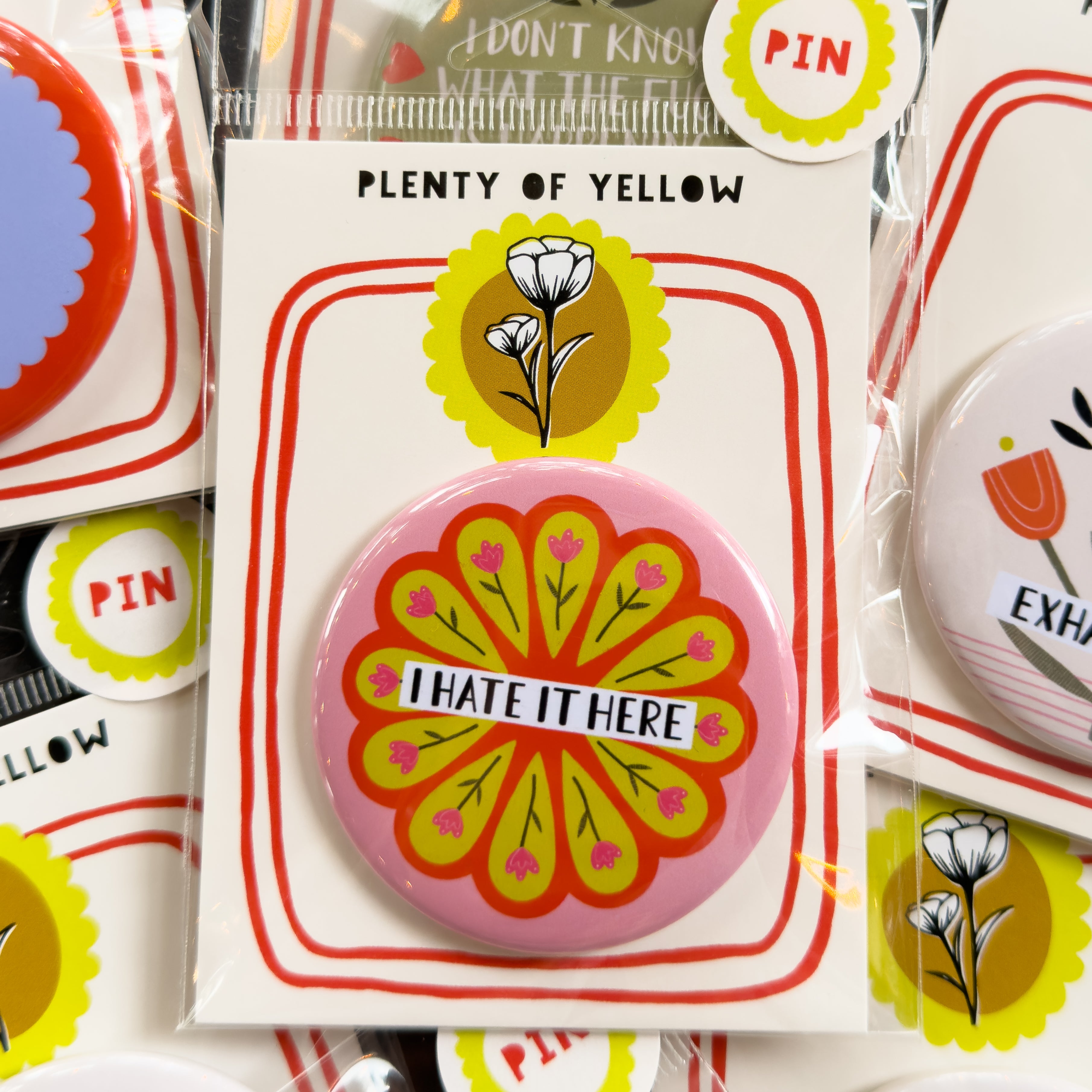 A round pinback button in clear plastic packaging, featuring a pink background with a circle of pink flowers on a yellow and red background and the text 'I HATE IT HERE'. The pin is mounted on a white backing card with red borders and a yellow logo, under the brand name 'PLENTY OF YELLOW'.
