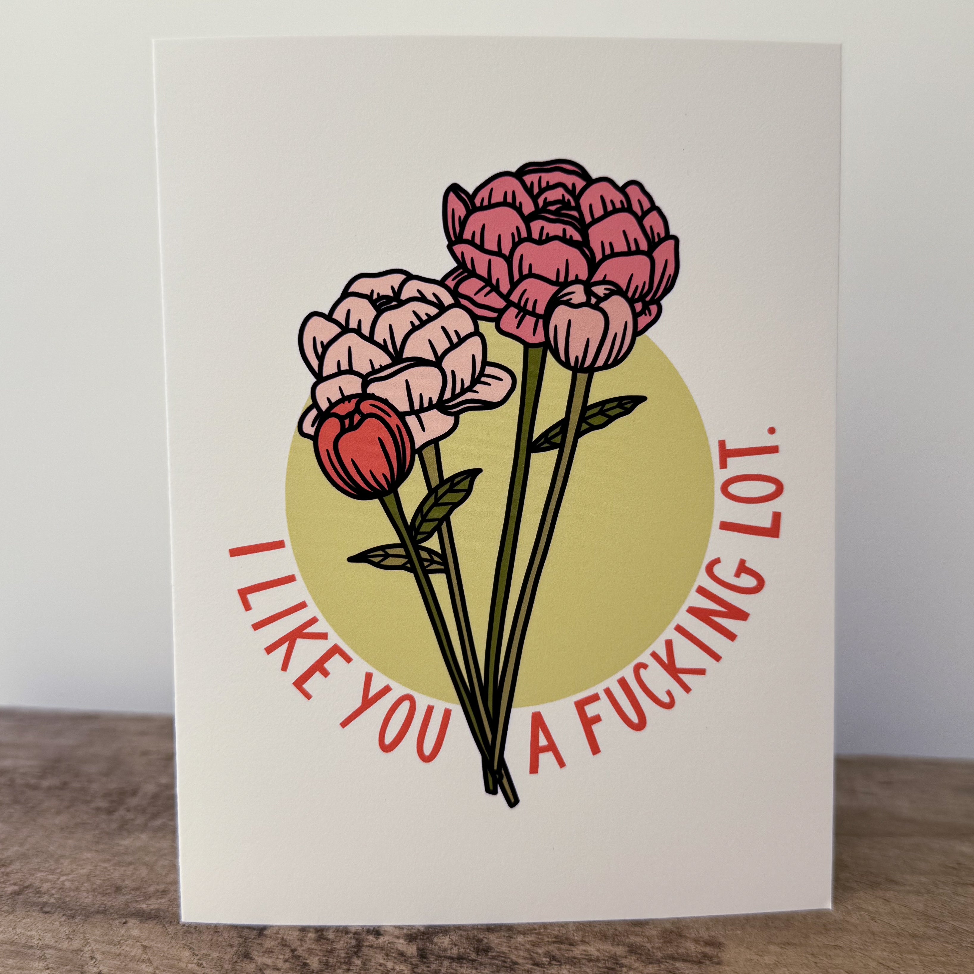 A greeting card with pink peony flowers and text 'I LIKE YOU A FUCKING LOT.' on a white background with a yellow circle.