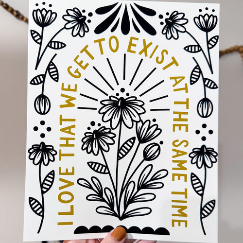 A hand holding an 8x10 print with flowers in the middle and around the edge with 'i love that we get to exist at the same time' written in an arch in between.