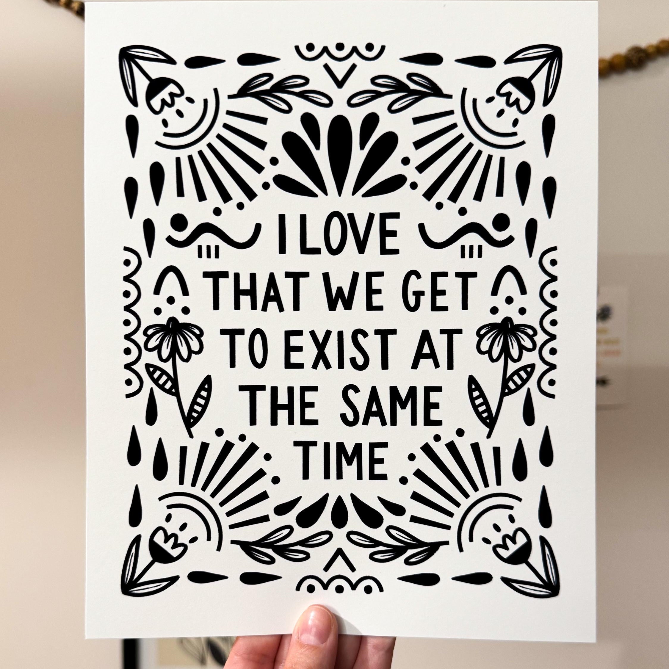 A hand holding an 8x10 art print that says 'I LOVE THAT WE GET TO EXIST AT THE SAME TIME' in black on white paper with floral and whimsy embellishments around it.