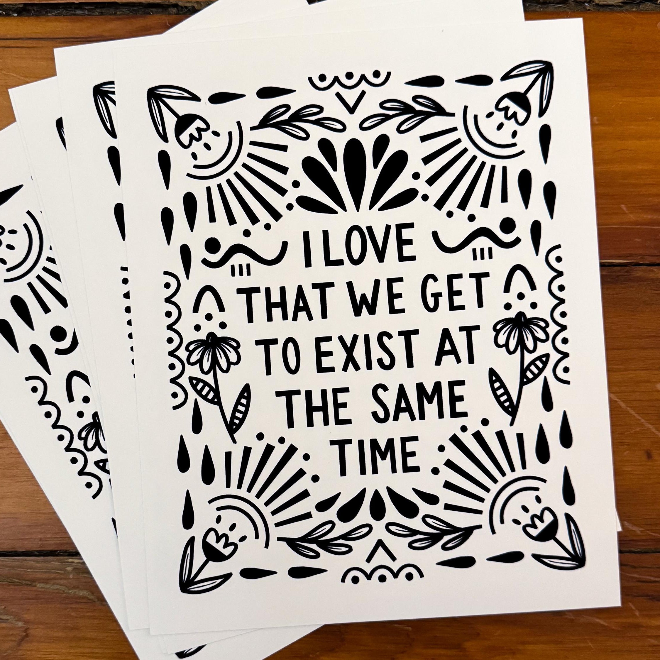 An 8x10 art print that says 'I LOVE THAT WE GET TO EXIST AT THE SAME TIME' in black on white paper with floral and whimsy embellishments around it.