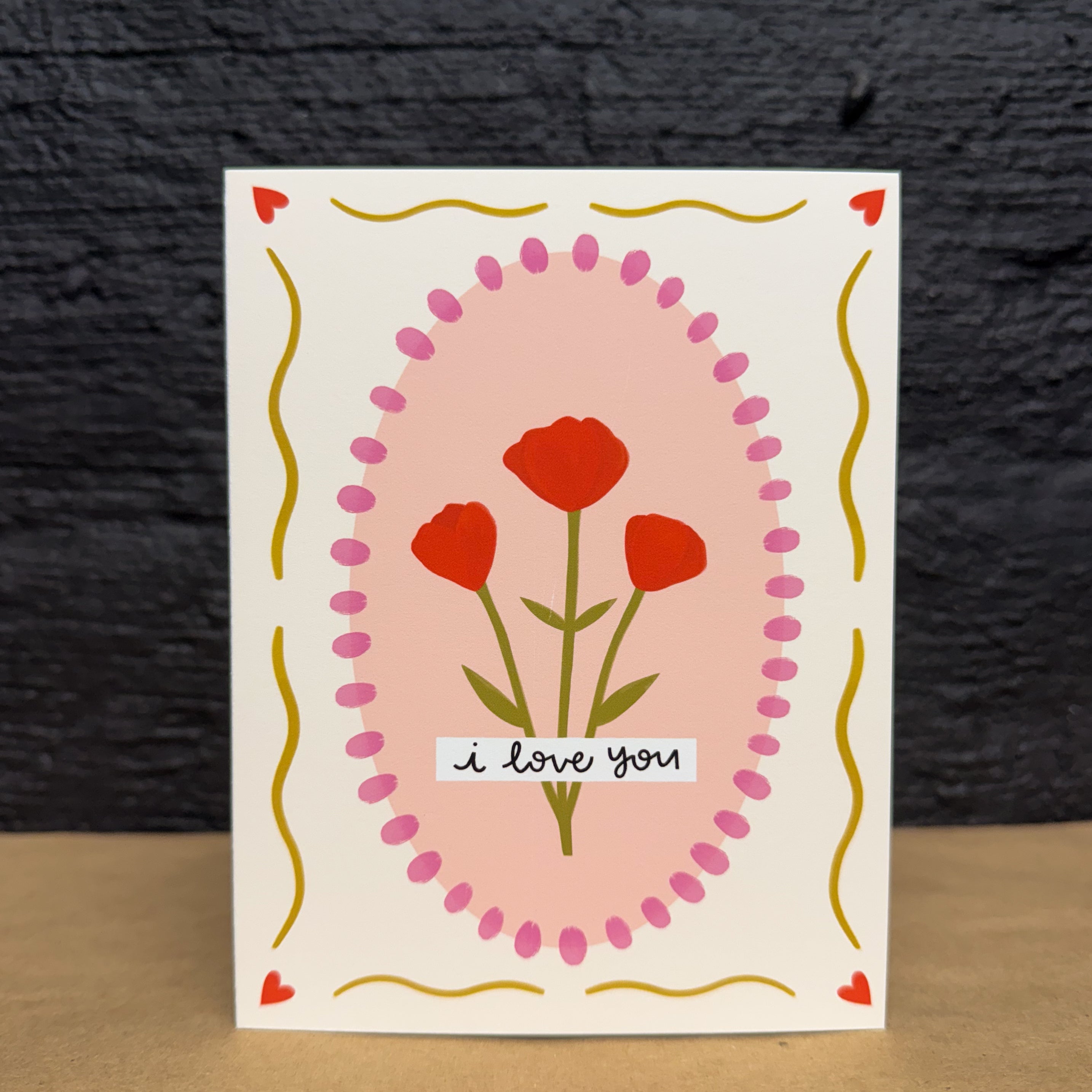 A greeting card that has a pink oval in the middle with flowers. On top of that, it says 'i love you'. There is a border of yellow wavy lines and red hearts.