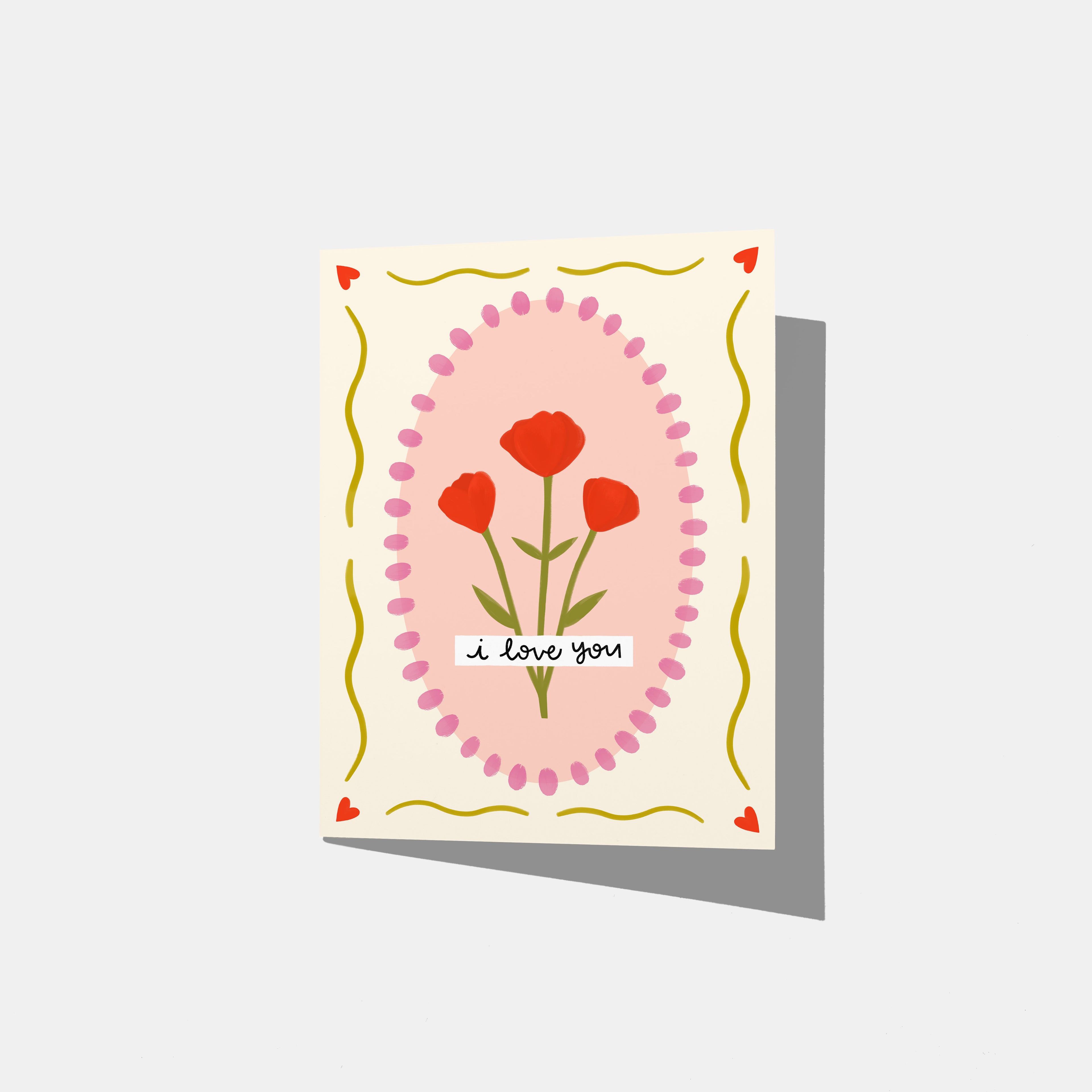 A mockup of a greeting card that has a pink oval in the middle with flowers. On top of that, it says 'i love you'. There is a border of yellow wavy lines and red hearts.