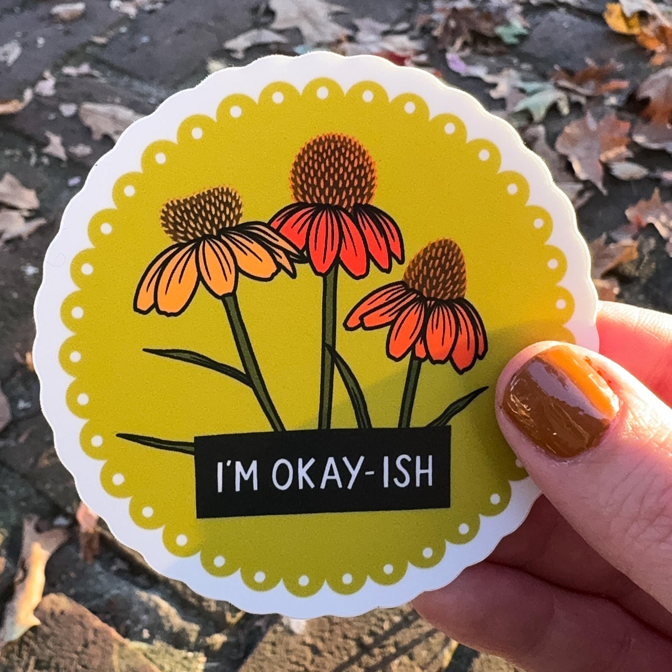 A hand holding a sticker with a floral design and 'I'm okay-ish' text on a yellow scalloped background.