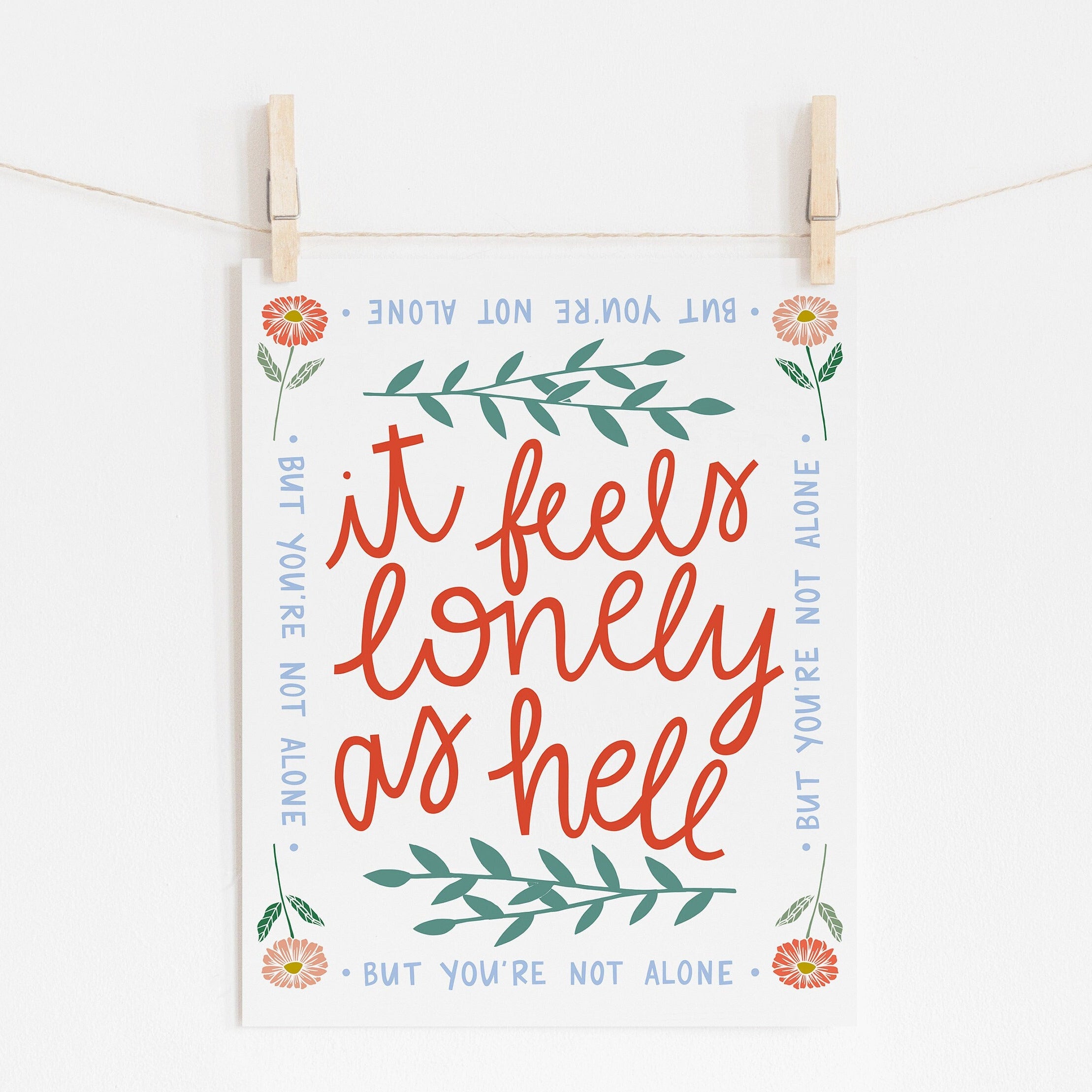 An 8x10 art print that says 'it feels lonely as hell' in the middle in red, surrounded by leaves. There is a border of flowers in each corner and it says 'but you're not alone' on each side.