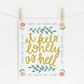 An 8x10 art print that says 'it feels lonely as hell' in the middle in yellow, surrounded by leaves. There is a border of flowers in each corner and it says 'but you're not alone' on each side.