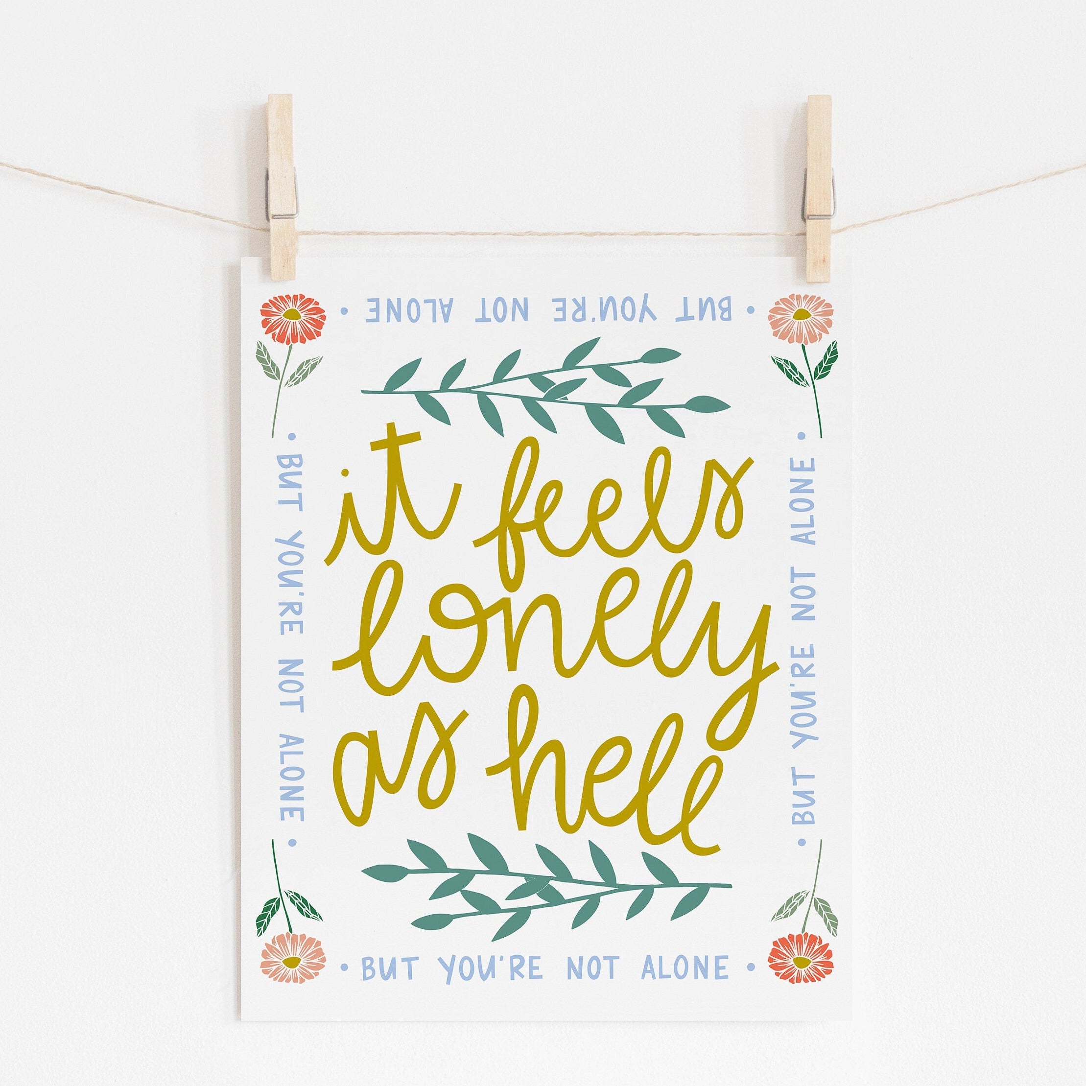 An 8x10 art print that says 'it feels lonely as hell' in the middle in yellow, surrounded by leaves. There is a border of flowers in each corner and it says 'but you're not alone' on each side.
