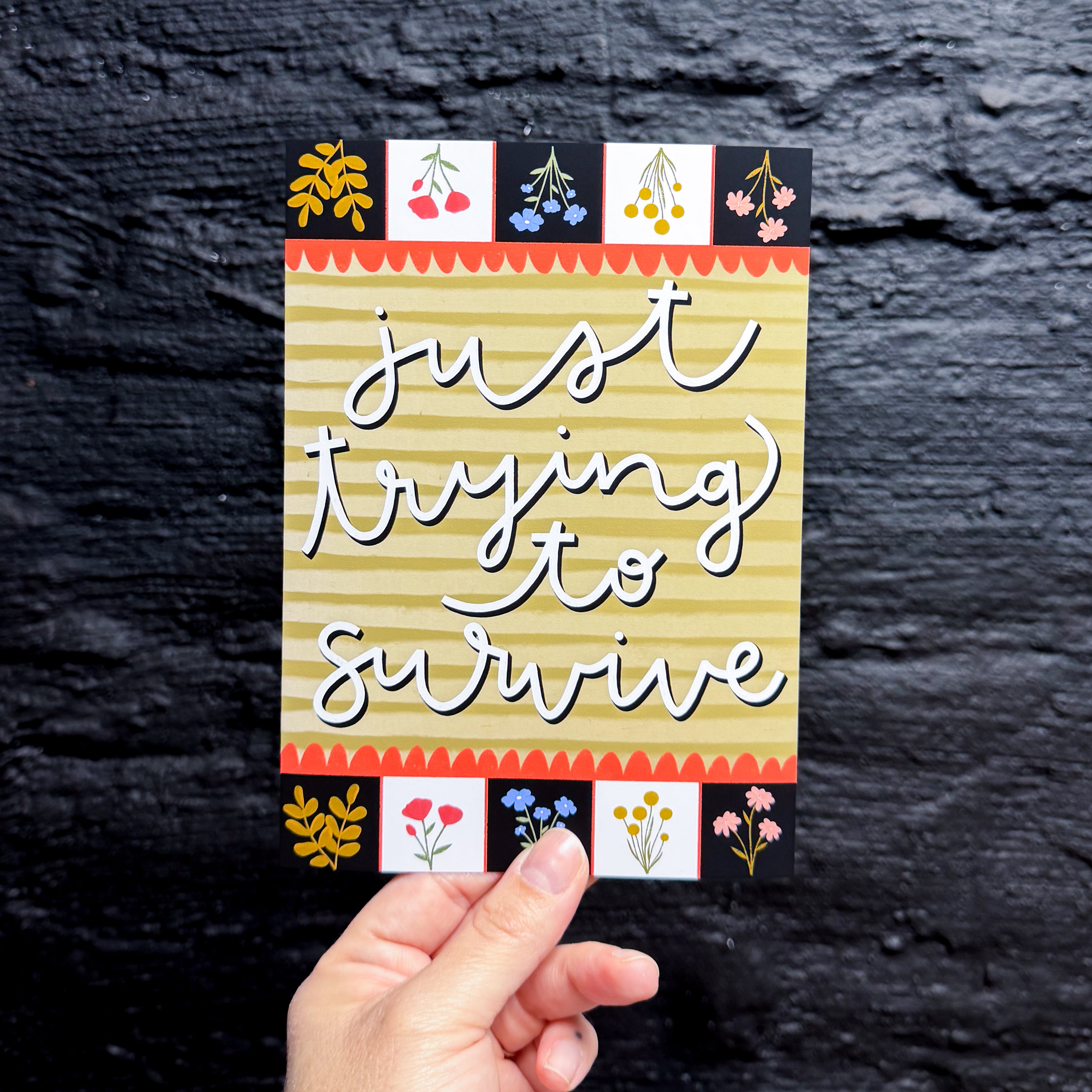 A hand holding a 5x7 print that says 'just trying to survive' in cursive on an olive green striped background. the top and bottom have black and white squares with flowers in them.