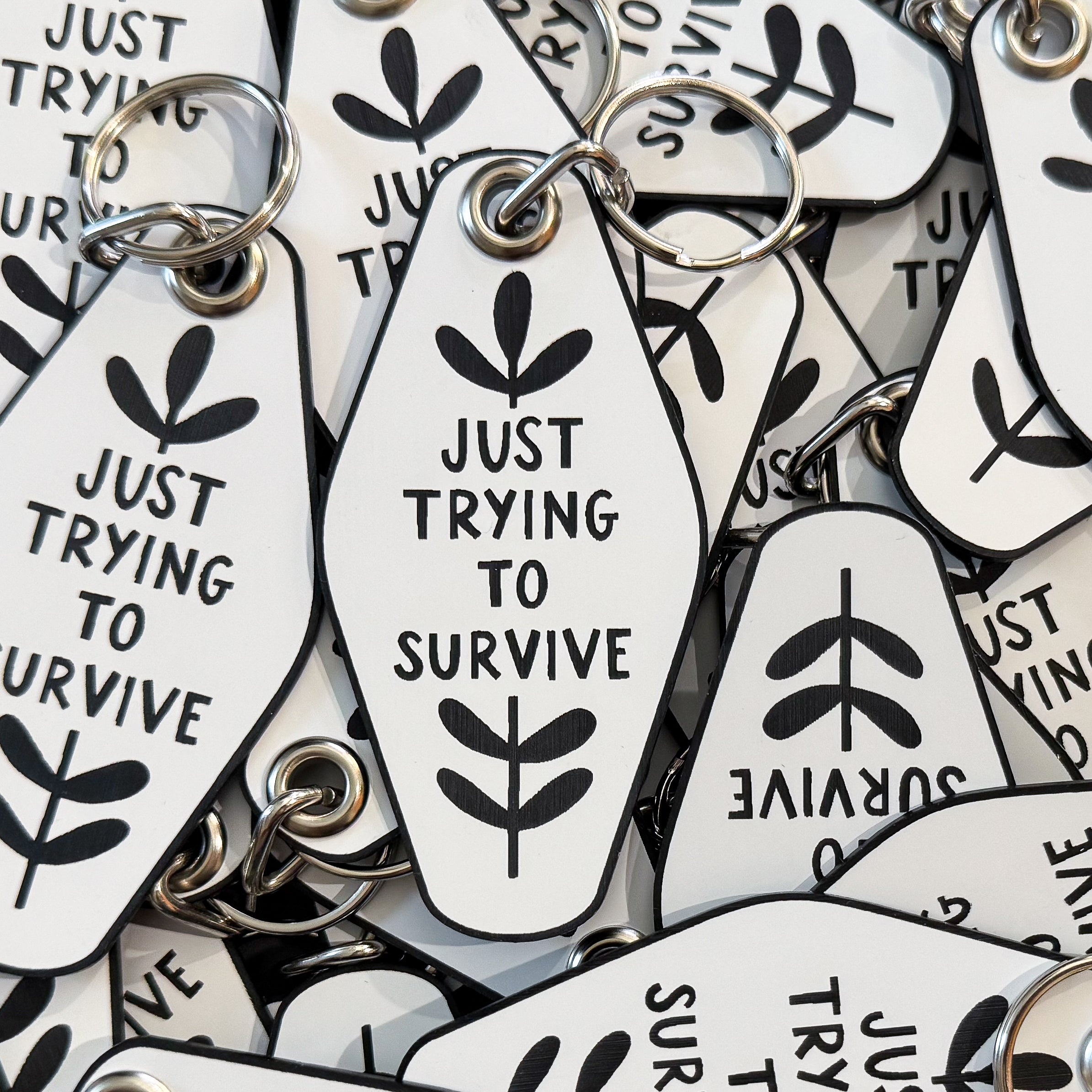 A pile of motel-style keychains in white with a black border that say 'JUST TRYING TO SURVIVE' in the middle surrounded by leaves.