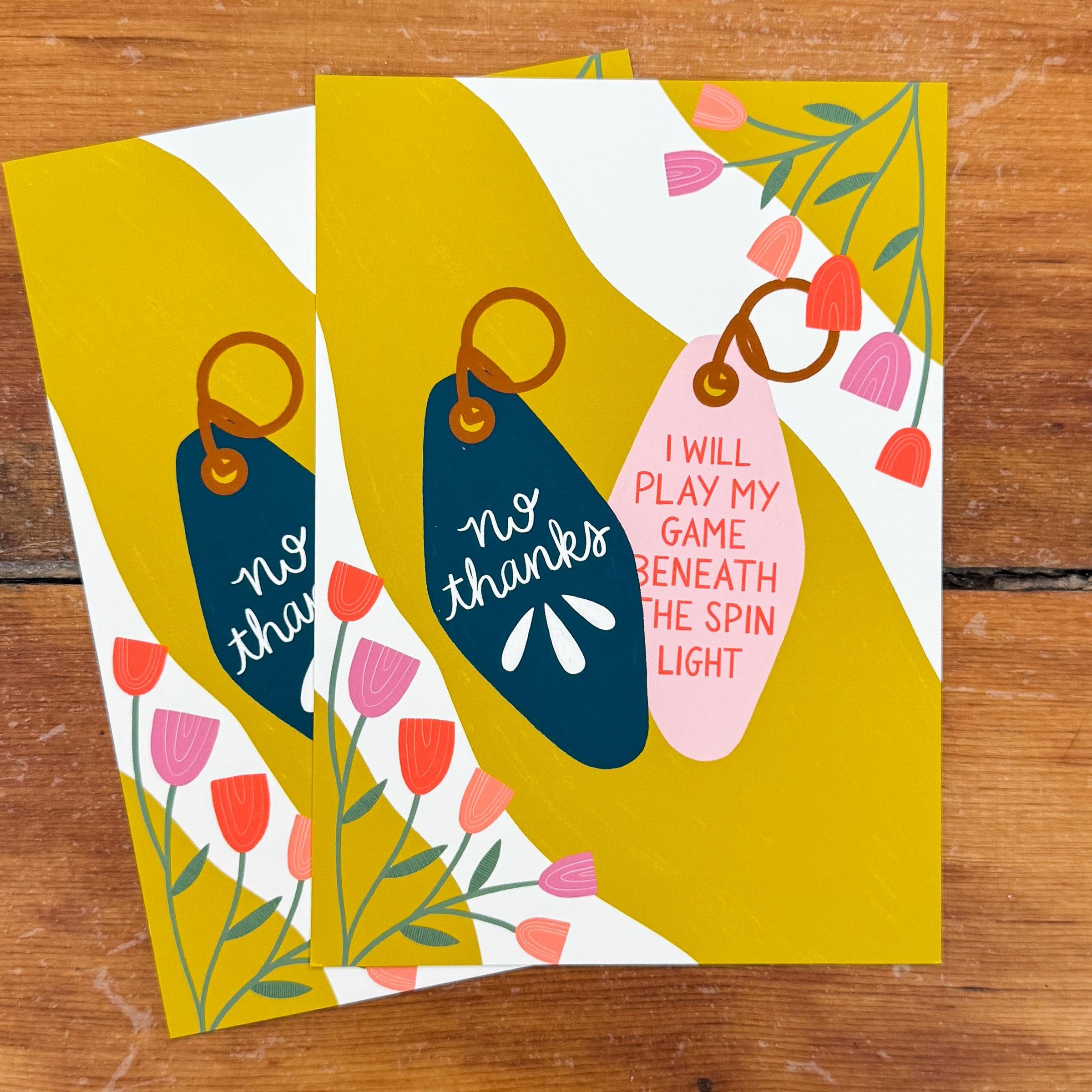 Two 5x7 art prints with two keychains. One keychain says 'no thanks' in off-white on a teal background. The other says 'I WILL PLAY MY GAME BENEATH THE SPIN LIGHT' in red on a pink background. They are on a wavy yellow and off-white background with floral embellishments in the corners.