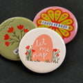 A round pinback button featuring a cream background with a peach colored grave surrounded by red flowers and the text 'let me nap'. The pin is surrounded by other pin designs.