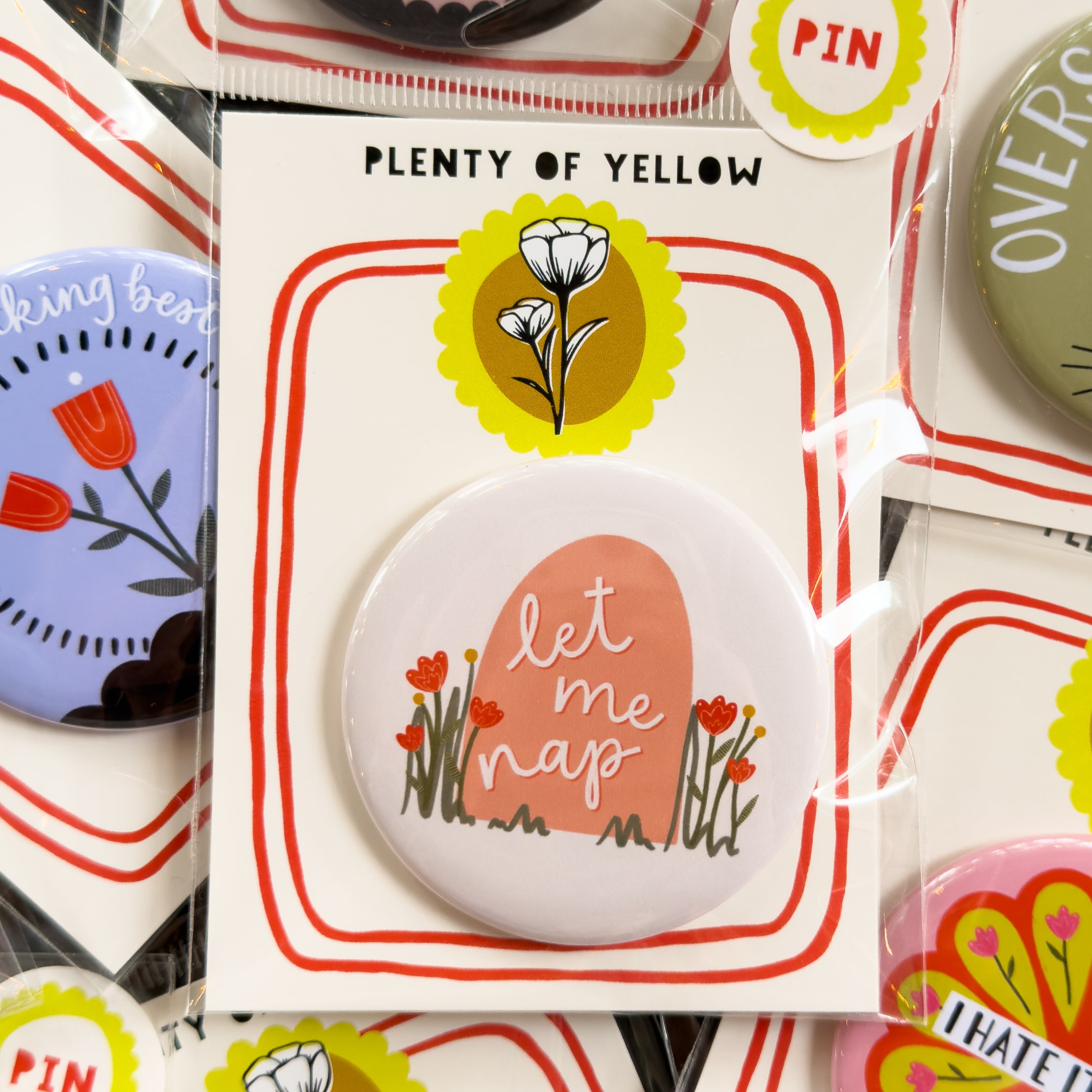 A round pinback button in clear plastic packaging, featuring a cream background with a peach colored grave surrounded by red flowers and the text 'let me nap'. The pin is mounted on a white backing card with red borders and a yellow logo, under the brand name 'PLENTY OF YELLOW'.