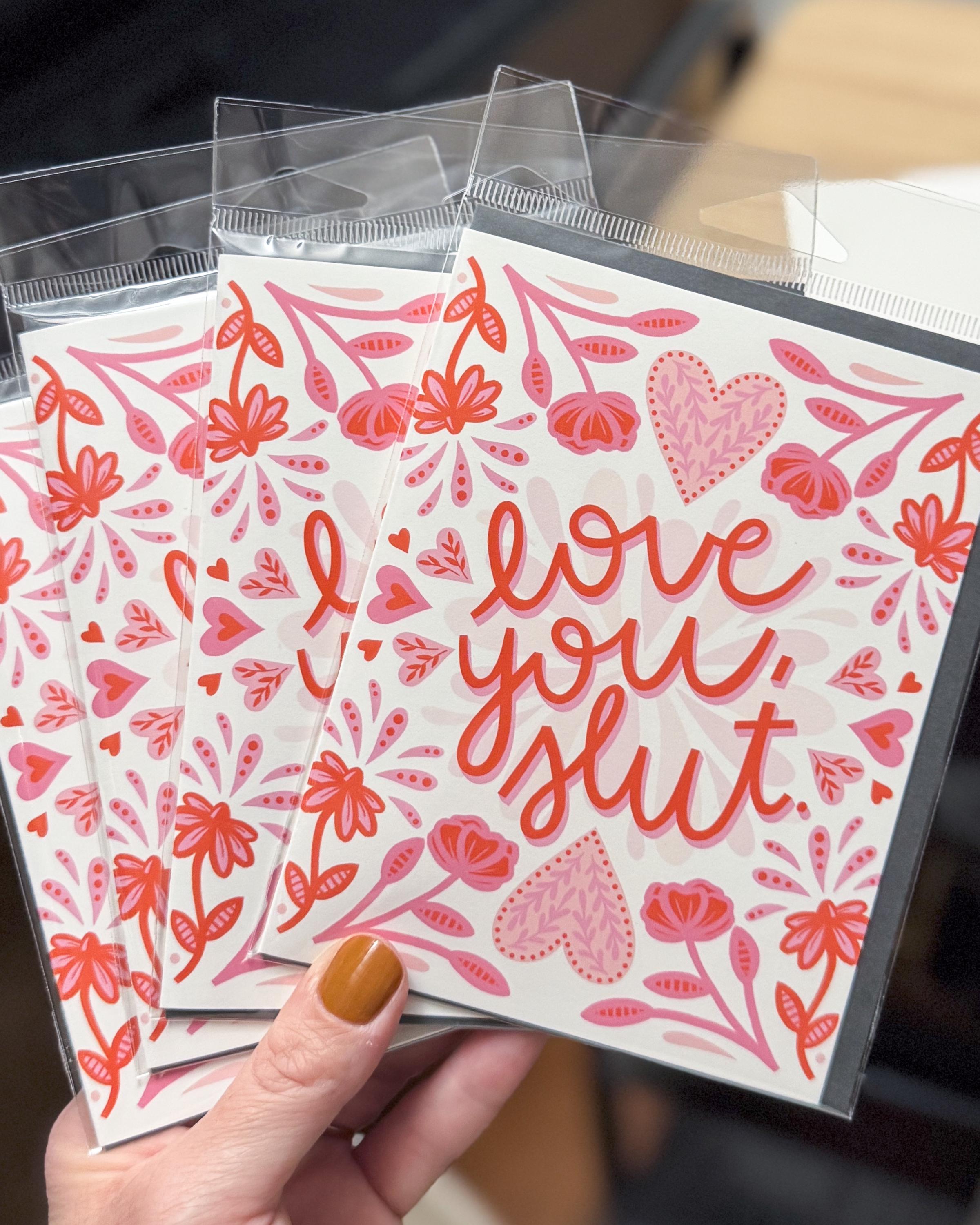 A hand holding a stack of greeting cards with 'love you, slut' text surrounded by floral red and pink patterns on a white background.