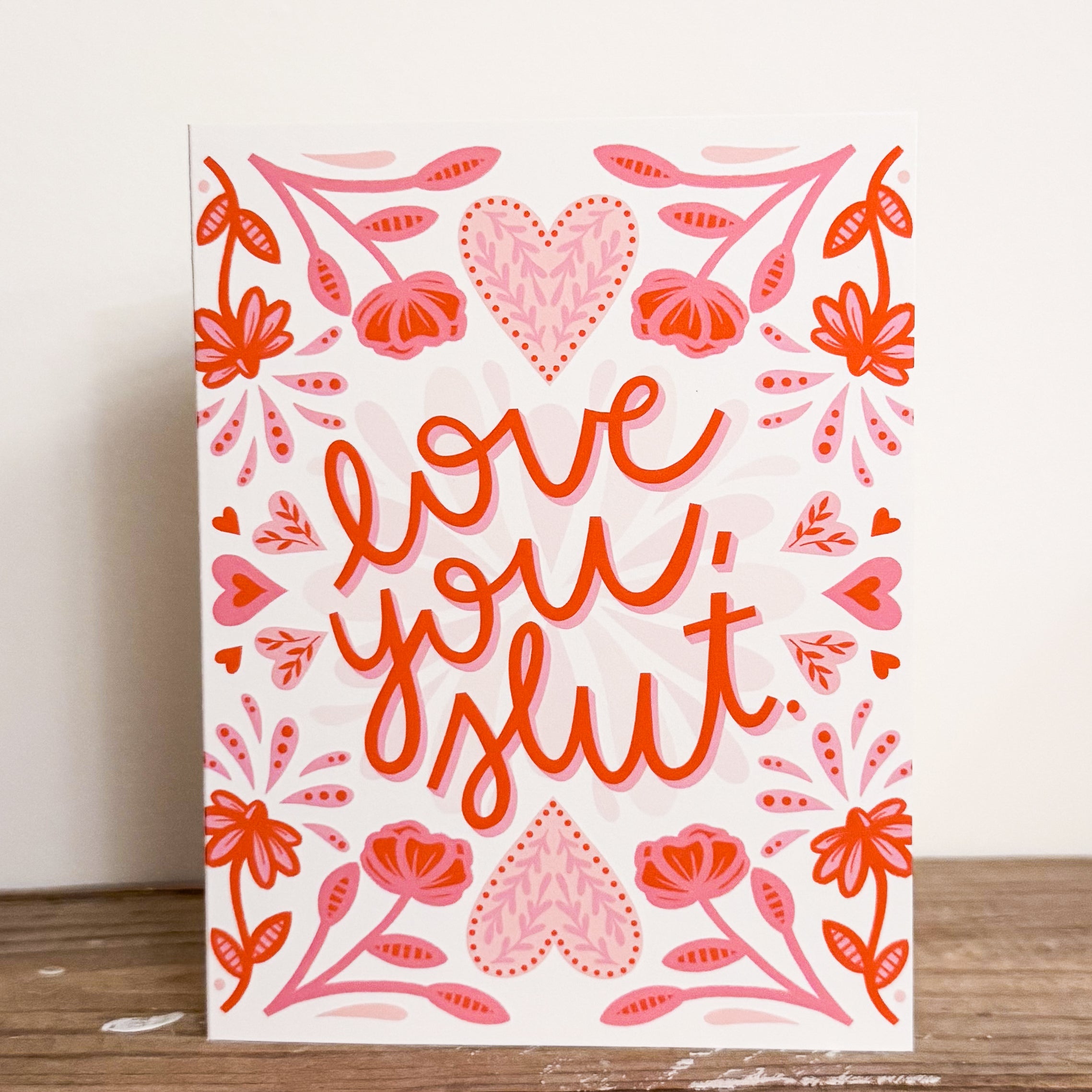 A greeting card with 'love you, slut' text surrounded by floral red and pink patterns on a white background.