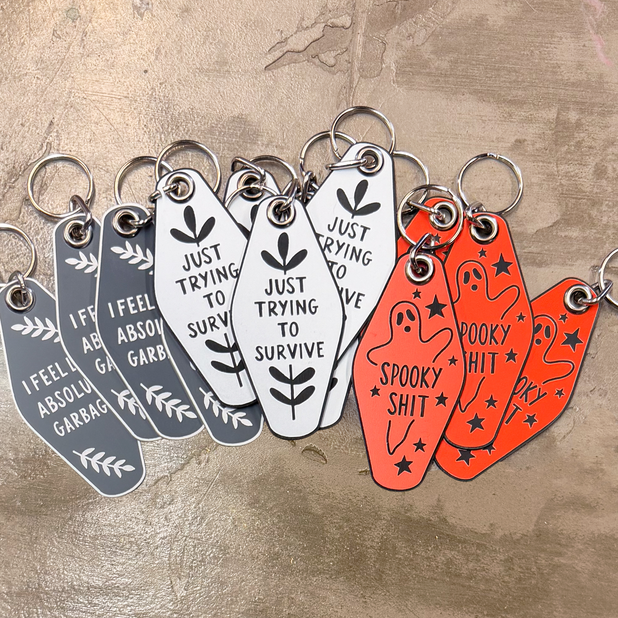 A pile of motel-style keychains in grey, white, and orange, with different sentiments on them: 'I FEEL LIKE ABSOLUTE GARBAGE', 'JUST TRYING TO SURVIVE', AND 'SPOOKY SHIT'.