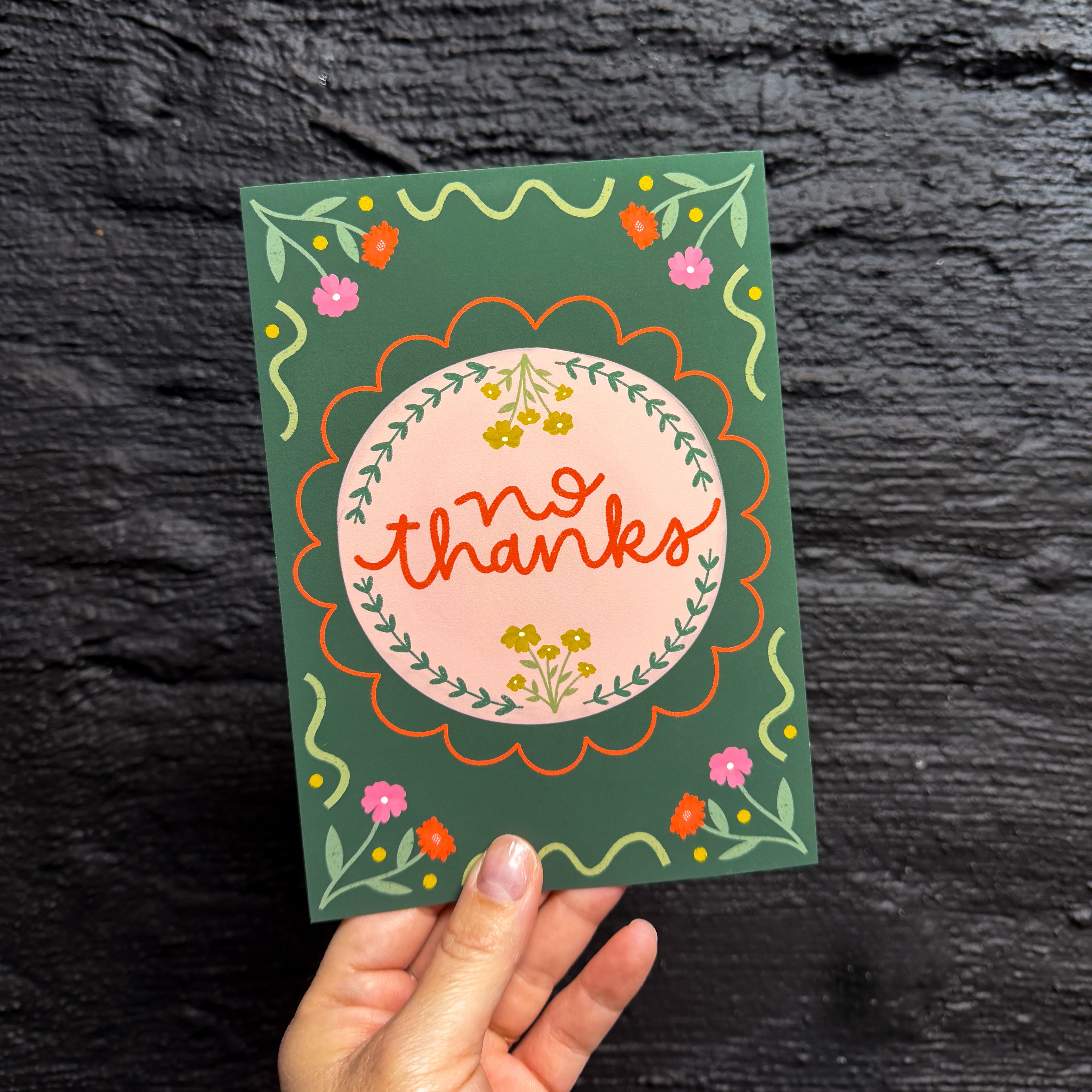 A hand holding a 5x7 art print with a dark green background that says 'no thanks' on a pink circle surrounded by embellishments and flowers.