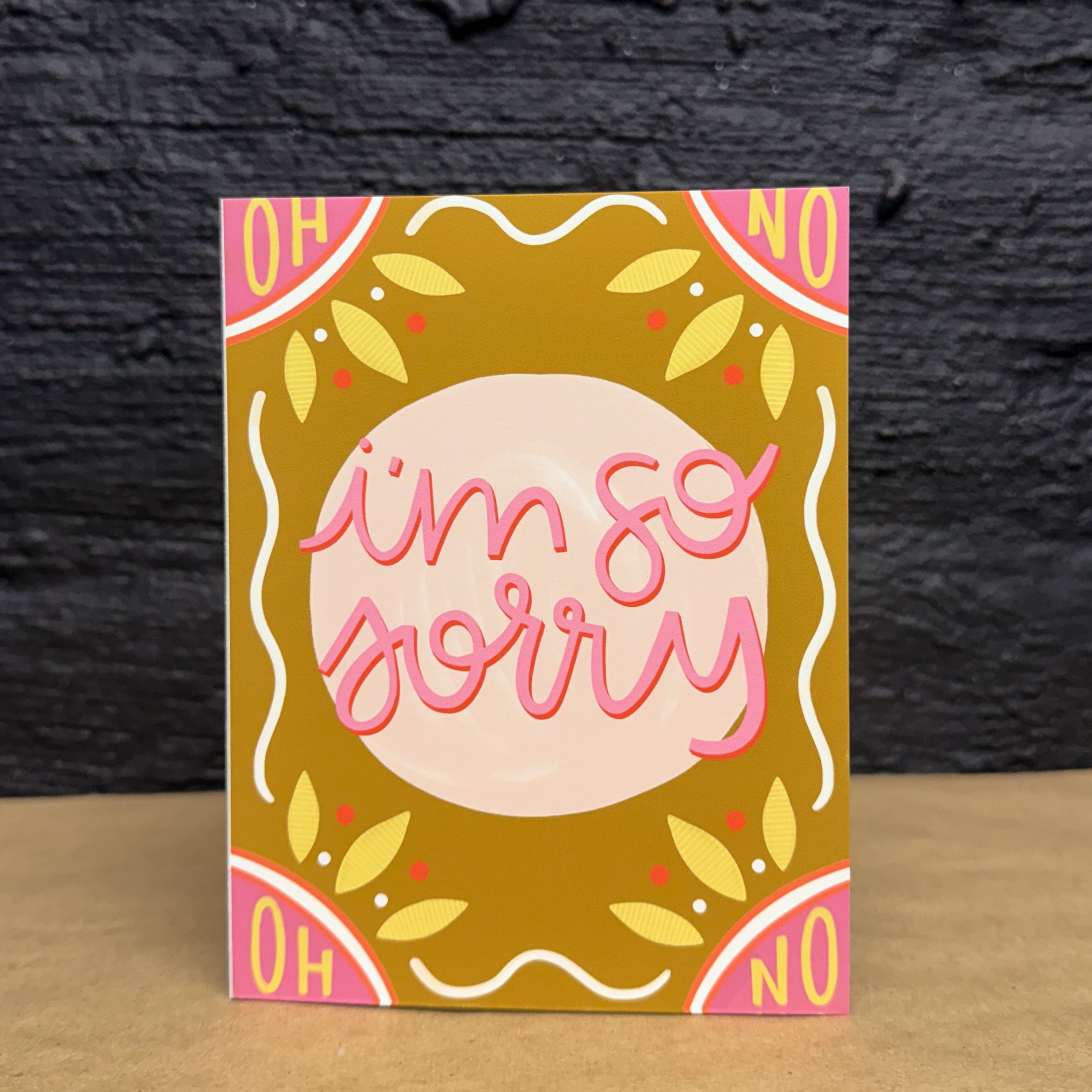 A dark yellow greeting card with a pink circle in the middle that says 'i'm so sorry' in pink and red on top. the top corners are pink and say 'oh' and 'no', as the same as the bottom. There are yellow and white embellishments around the card.