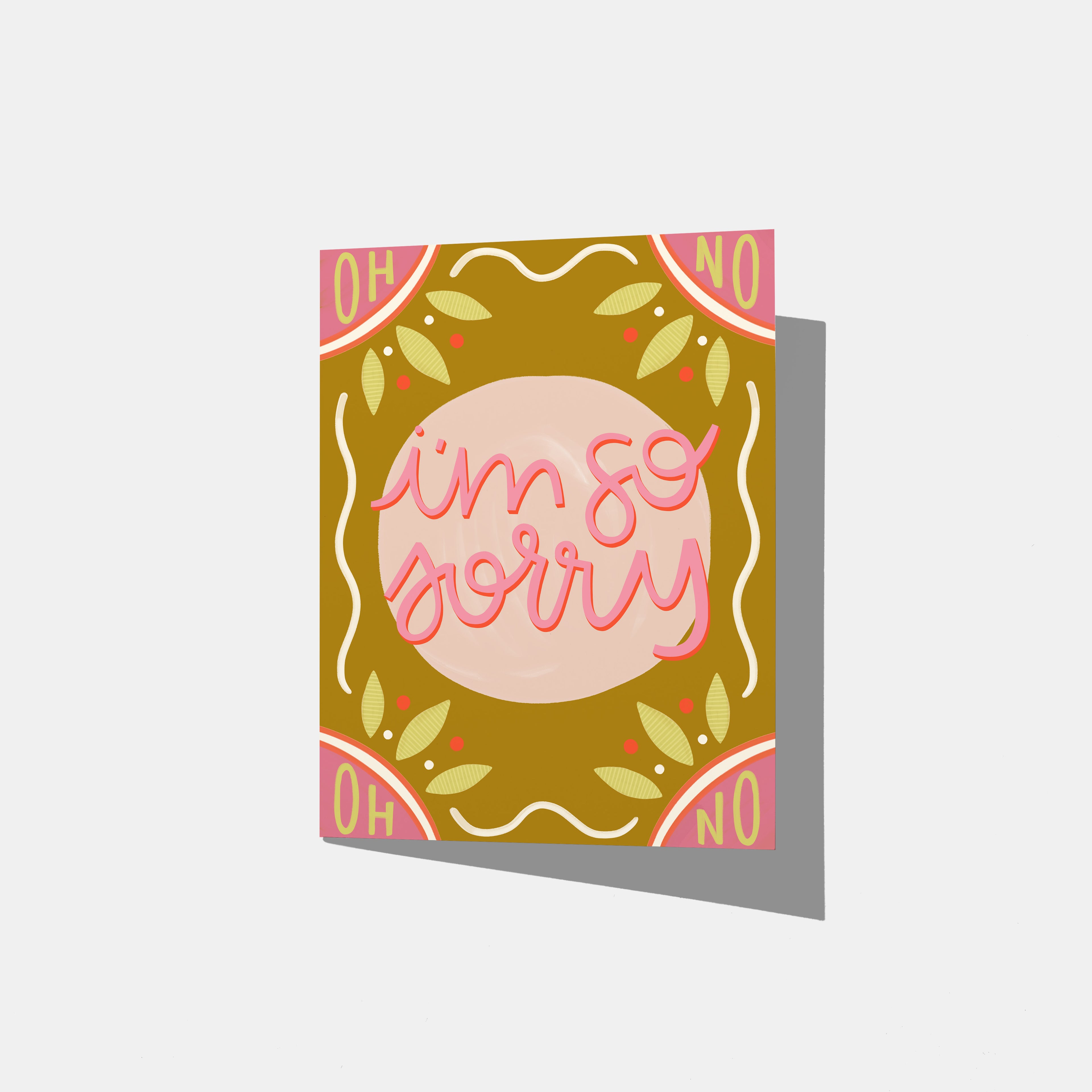 A mockup of a dark yellow greeting card with a pink circle in the middle that says 'i'm so sorry' in pink and red on top. the top corners are pink and say 'oh' and 'no', as the same as the bottom. There are yellow and white embellishments around the card.