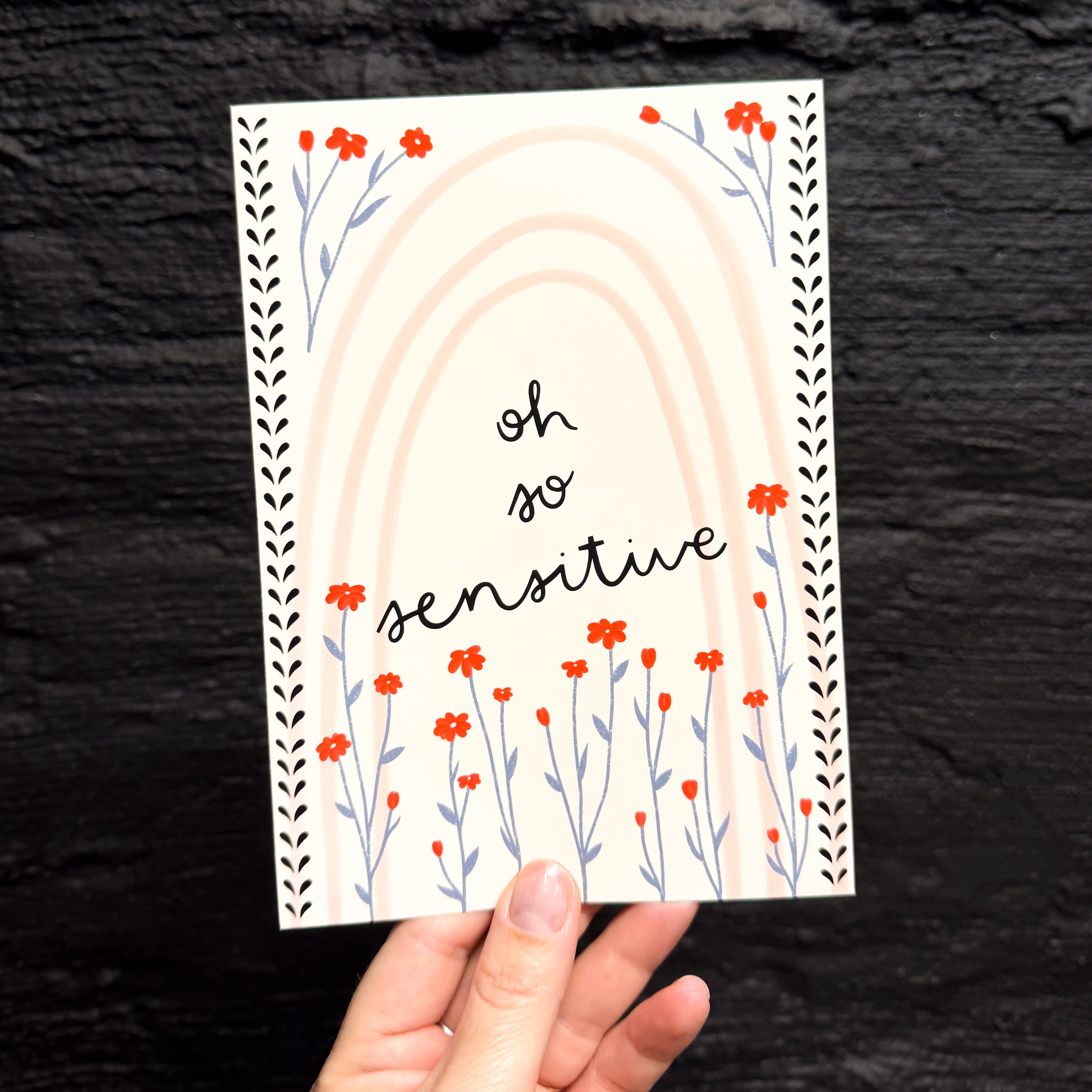 A hand holding a 5x7 art print that says 'oh so sensitive' in the middle, surrounded by red and blue flowers. There is a pink rainbow in the background with black flourishes down the length of the sides.