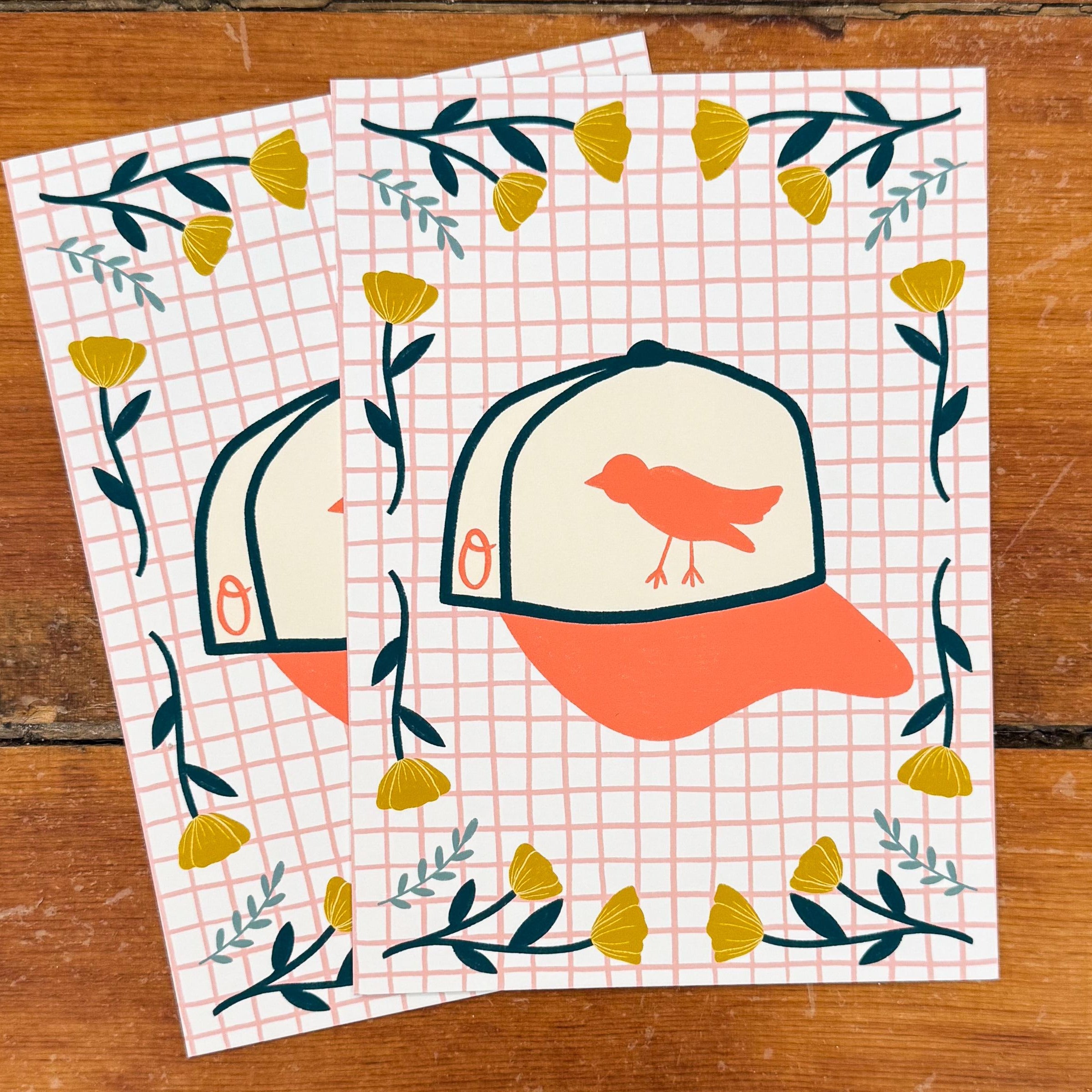Two 5x7 art prints of an Orioles baseball hat on a pink grid background surrounded by yellow and teal flowers.