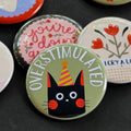 A round pinback button featuring a sage green background with a black cat in a yellow and pink party hat. The text ‘OVERSTIMULATED’ sits across the top. The pin is surrounded by other pin designs.