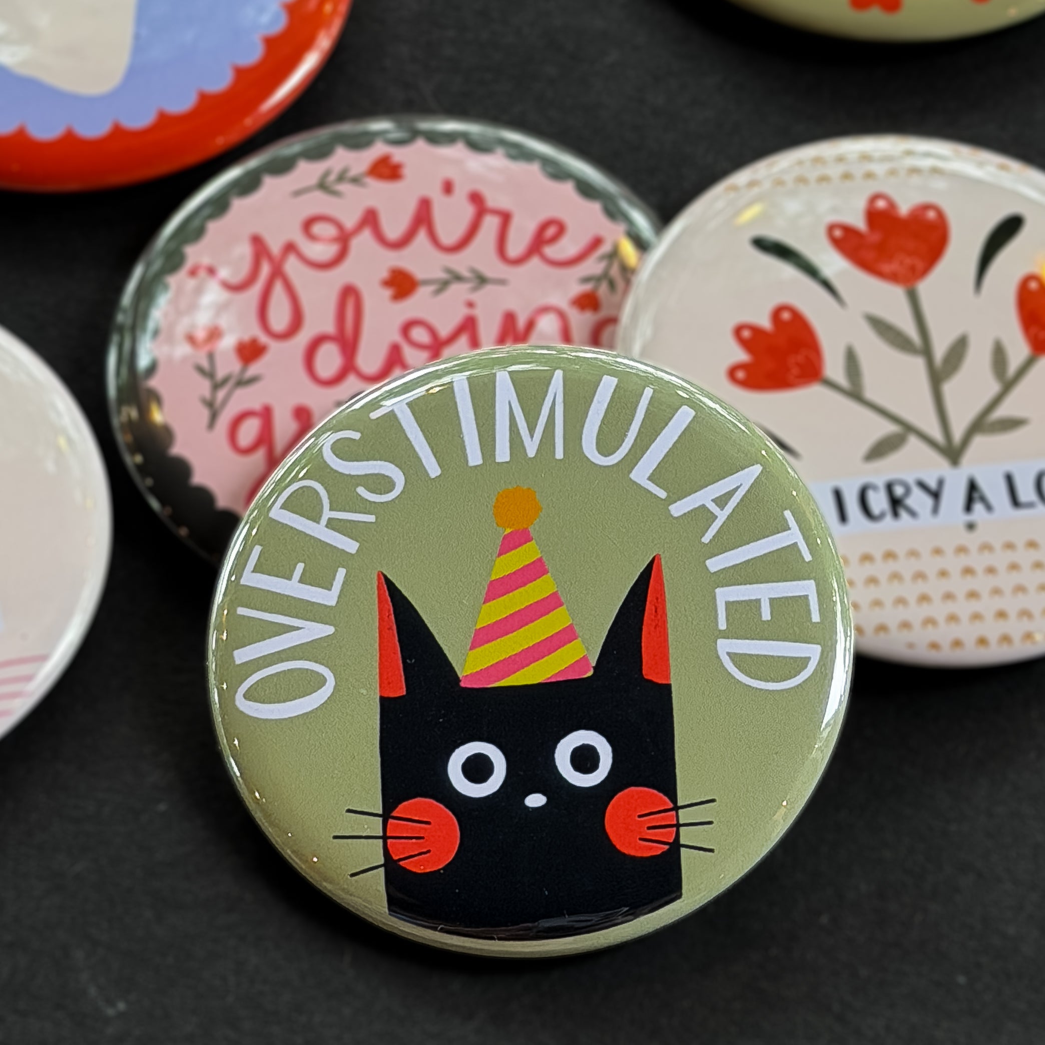 A round pinback button featuring a sage green background with a black cat in a yellow and pink party hat. The text ‘OVERSTIMULATED’ sits across the top. The pin is surrounded by other pin designs.