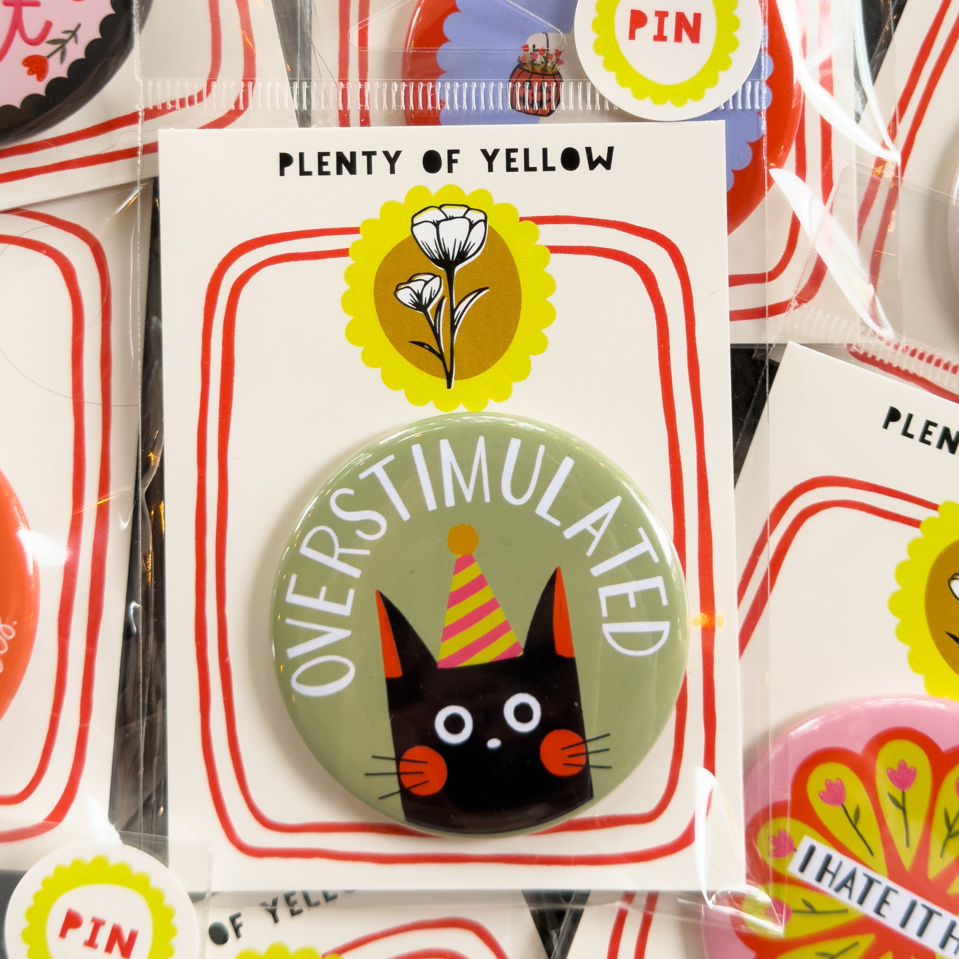 A round pinback button in clear plastic packaging, featuring a sage green background with a black cat in a yellow and pink party hat. The text ‘OVERSTIMULATED’ sits across the top. The pin is mounted on a white backing card with red borders and a yellow logo, under the brand name 'PLENTY OF YELLOW'.