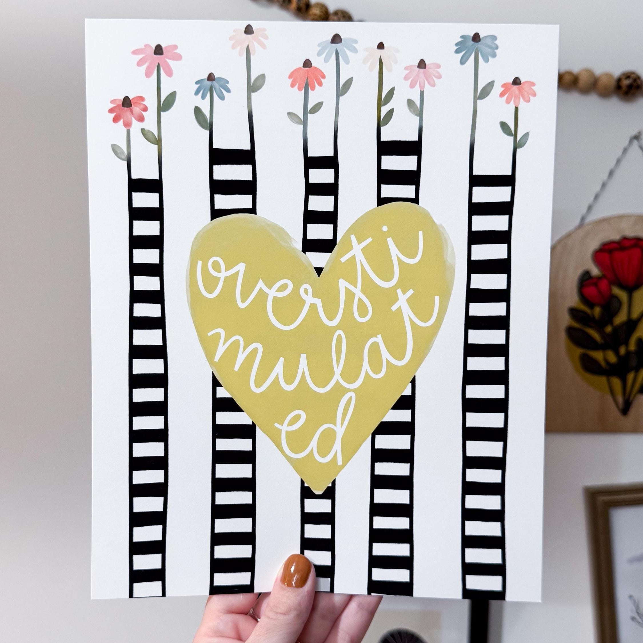 A hand holding an 8x10 print with a yellow heart in the middle that says 'overstimulated'. The background has flowers coming from black and white striped stems.