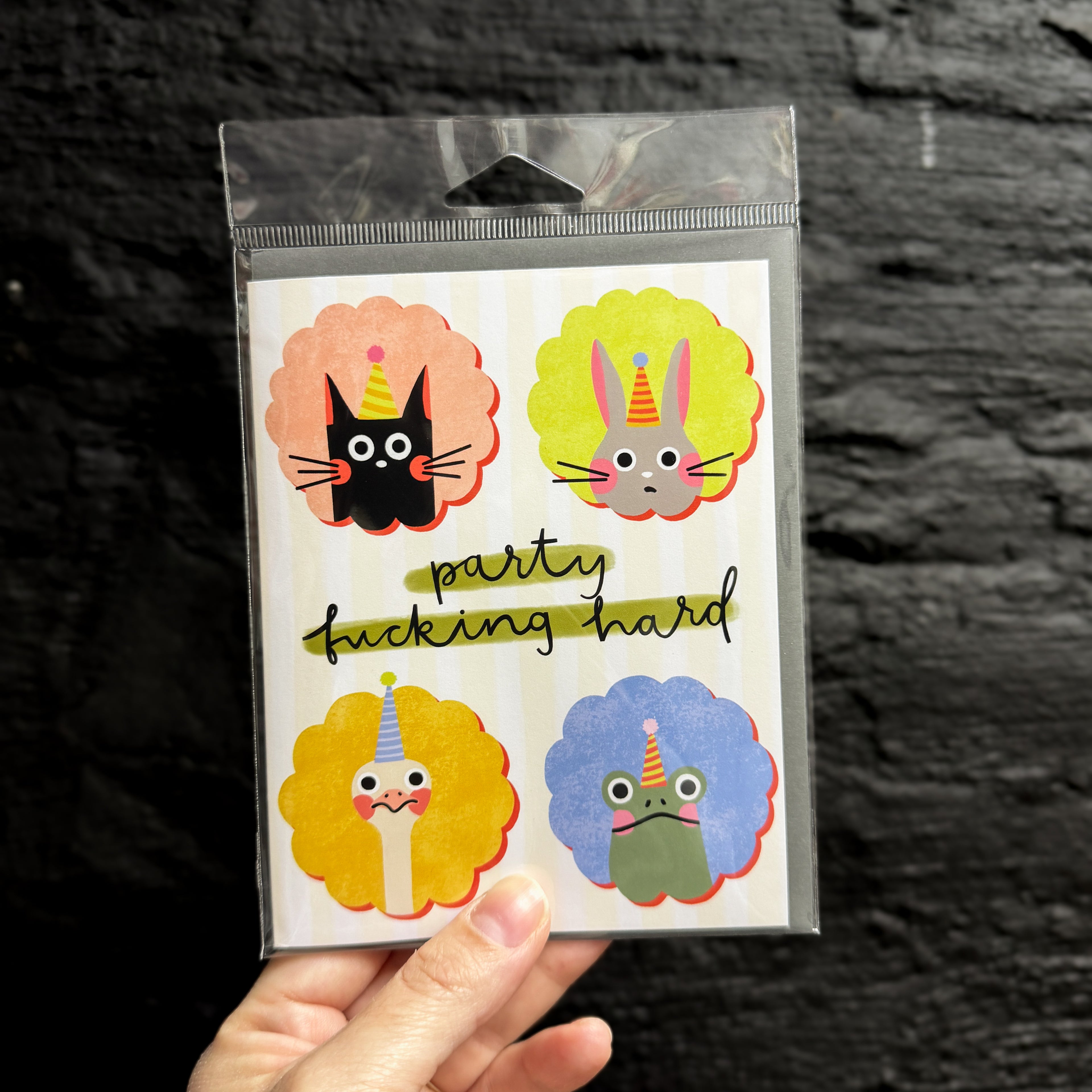 A hand holding a greeting card with that says 'party fucking hard' in the middle surrounded by four portraits of animals (a cat, a bunny, an ostrich, and a frog) in party hats looking unamused.