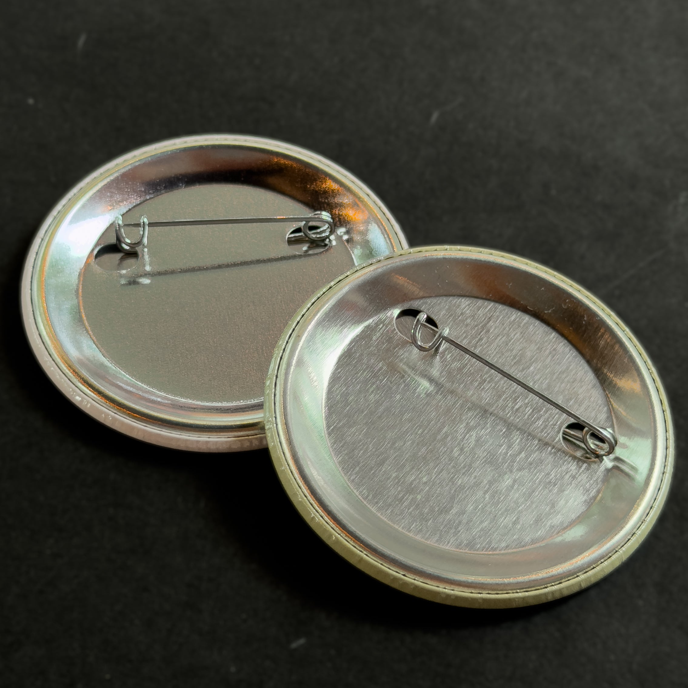 Two round silver pinback buttons showing their backs, with safety pins attached, on a dark background.