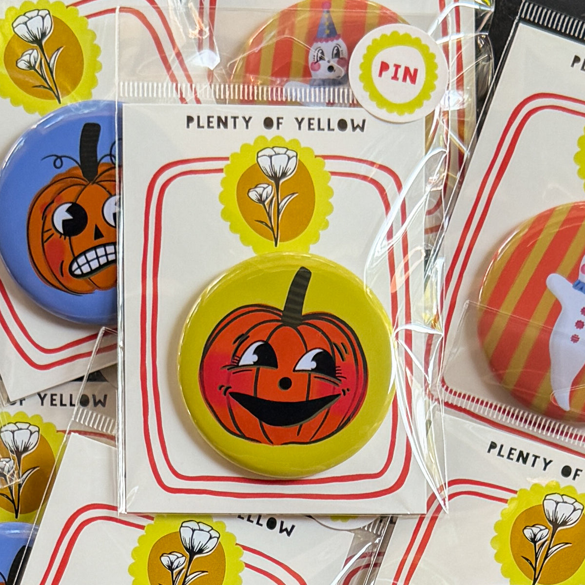 A round pinback button in clear plastic packaging, featuring a happy orange pumpkin named Clementine with a yellow background. The pin is mounted on a white backing card with red borders and a yellow logo, under the brand name 'PLENTY OF YELLOW'.