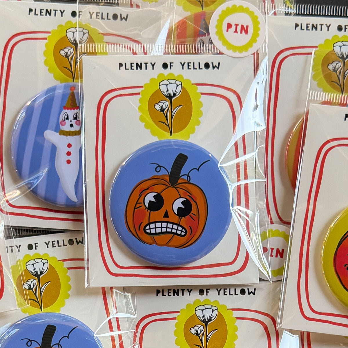 A round pinback button in clear plastic packaging, featuring a nervous orange pumpkin named Theo with a blue background. The pin is mounted on a white backing card with red borders and a yellow logo, under the brand name 'PLENTY OF YELLOW'.