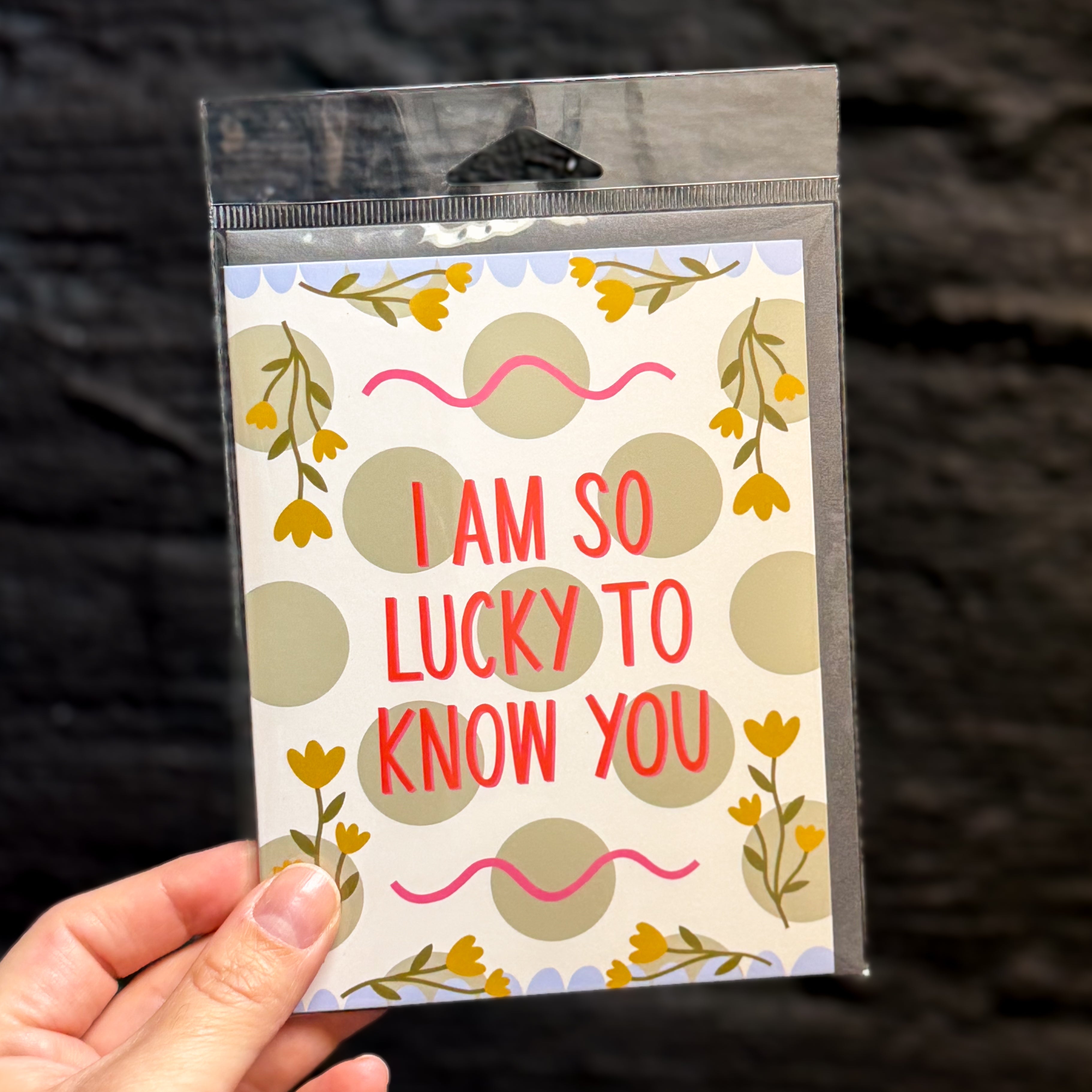 A hand holding a packaged greeting card against a dark textured background. The card has a white base with sage green polka dots, yellow floral sprigs, pink squiggly lines, and red text that reads, 'I AM SO LUCKY TO KNOW YOU'.