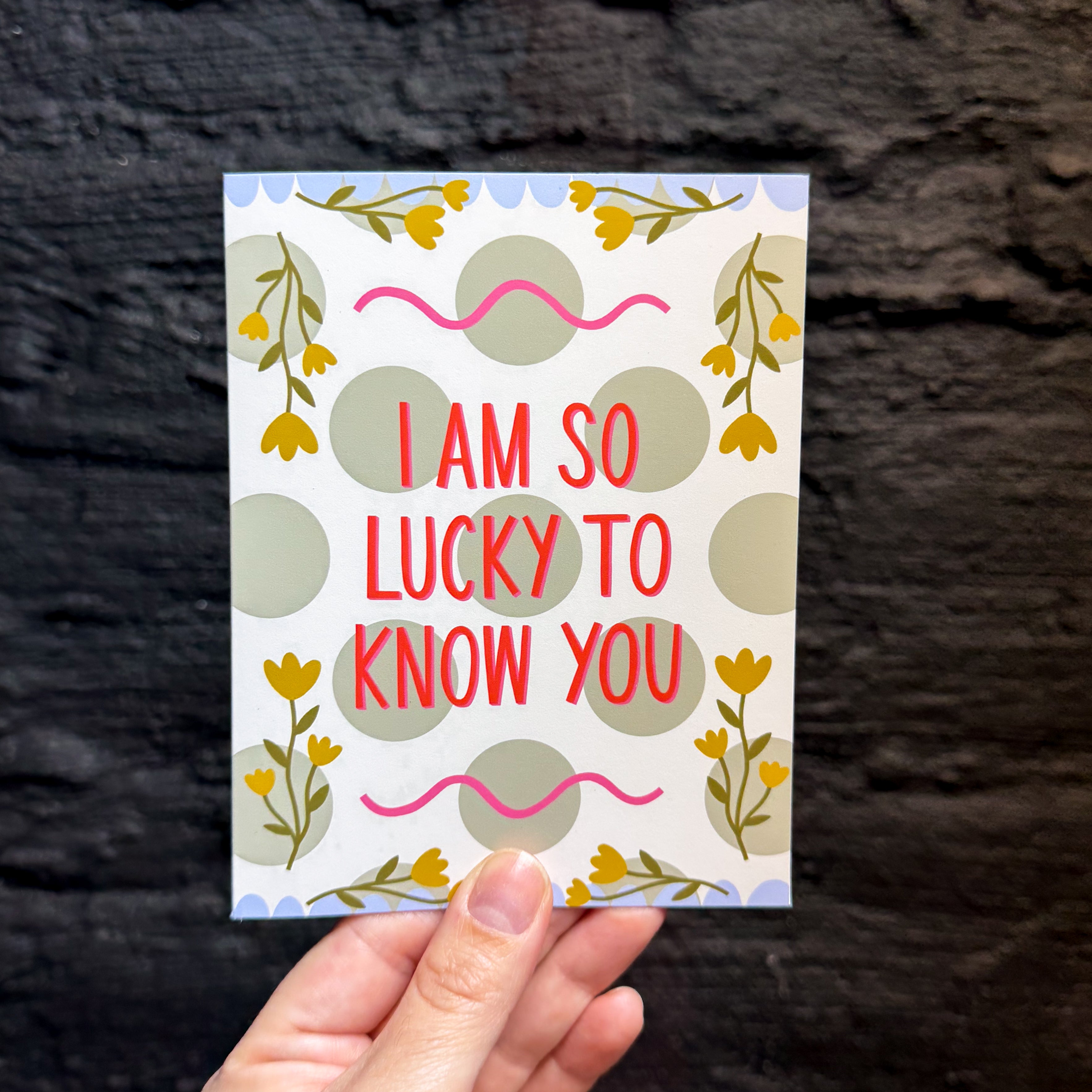 A hand holding a greeting card against a dark textured background. The card has a white base with sage green polka dots, yellow floral sprigs, pink squiggly lines, and red text that reads, 'I AM SO LUCKY TO KNOW YOU'.