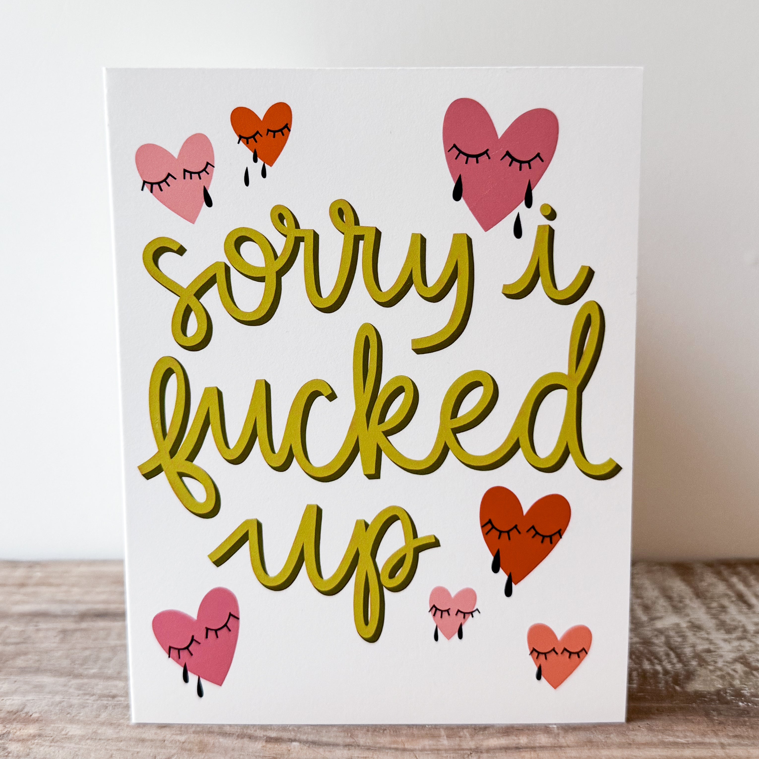 A greeting card with green text 'sorry i fucked up' in a yellow/green with red and pink crying heart illustrations on a white background.
