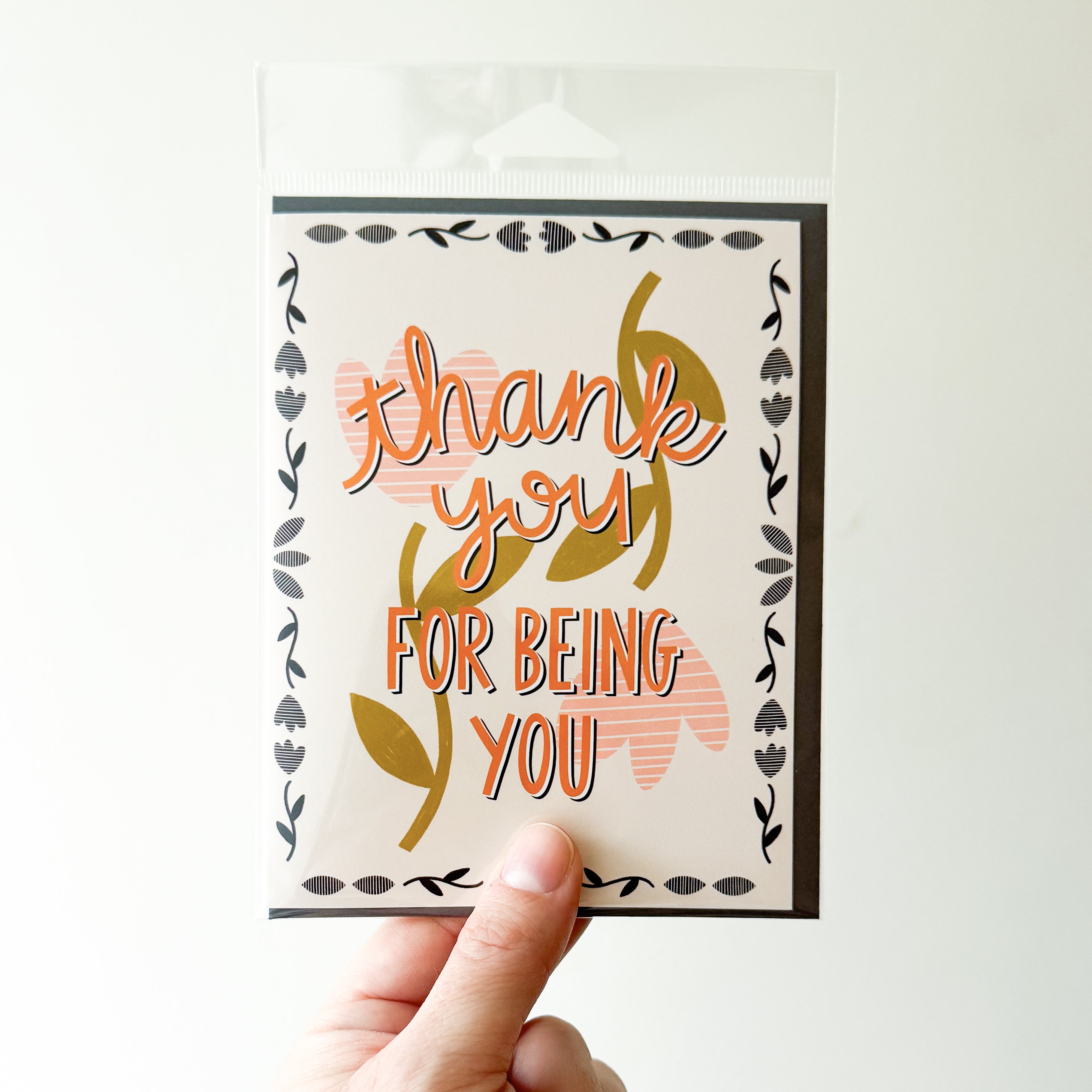 A hand holding a greeting card with 'thank you FOR BEING YOU' in orange text with a peach flower background and border of little black flowers.