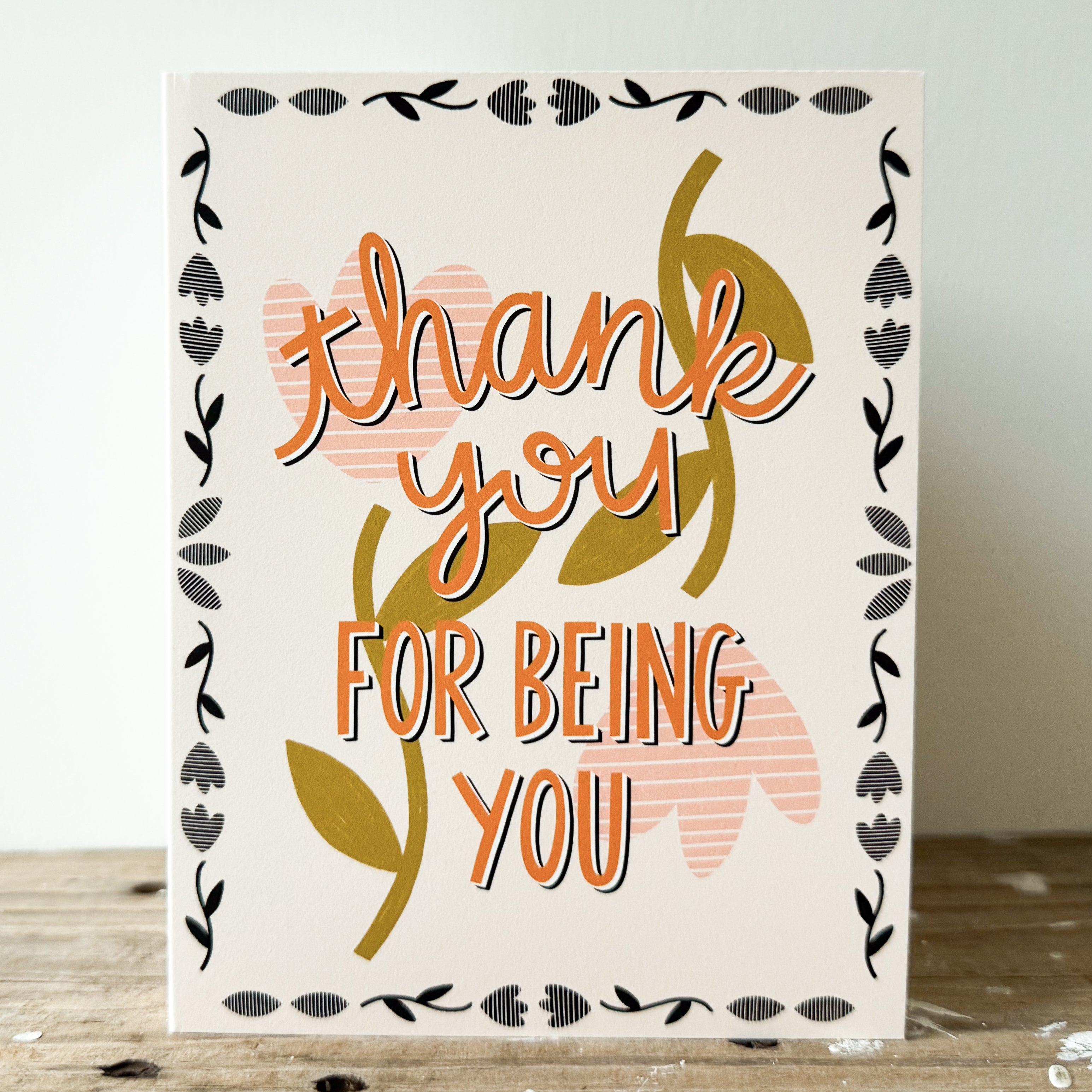 A greeting card with 'thank you FOR BEING YOU' in orange text with a peach flower background and border of little black flowers.