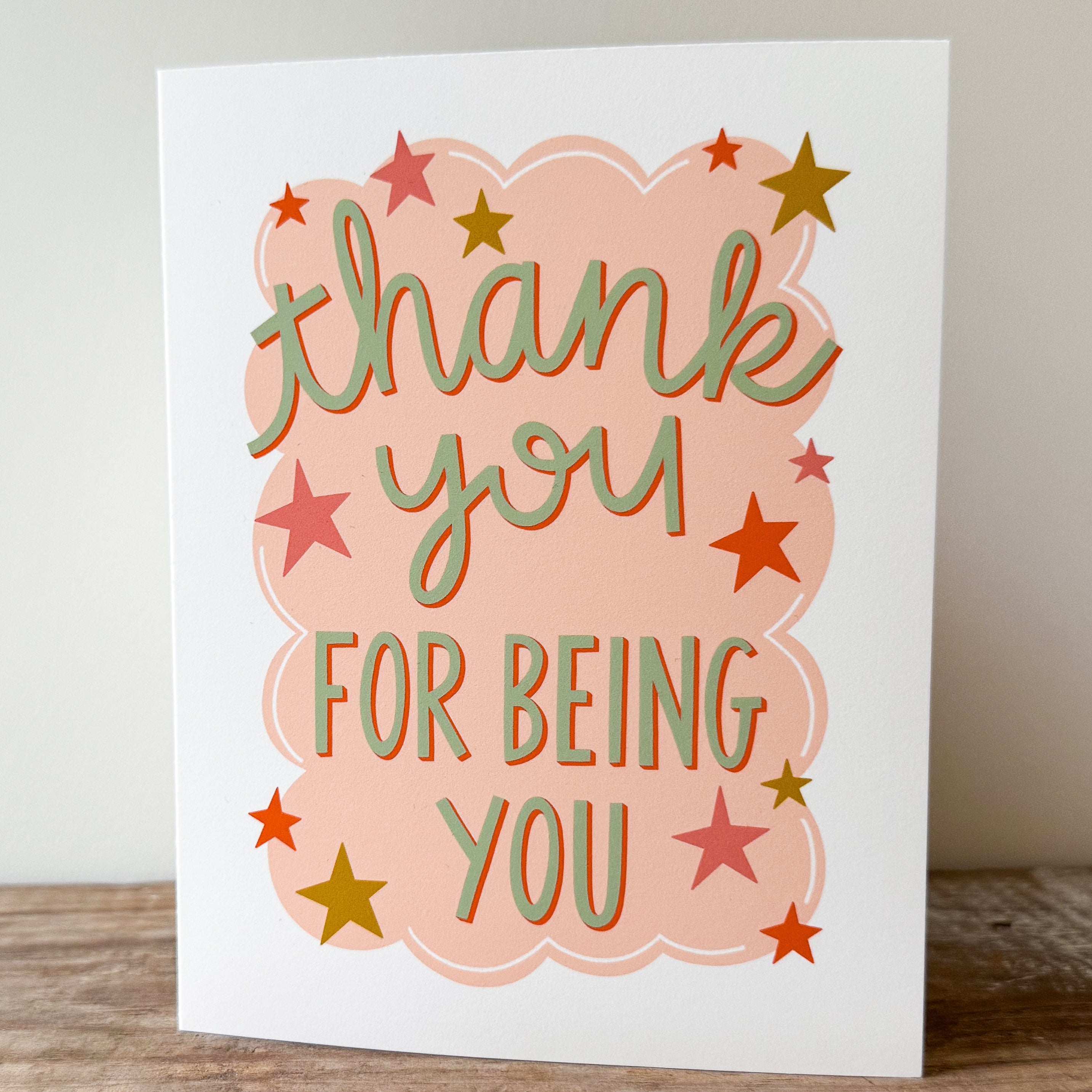 A greeting card with 'thank you for being you' text in green with a red drop shadow and star decorations on a  peach cloud-like shape on a white background.