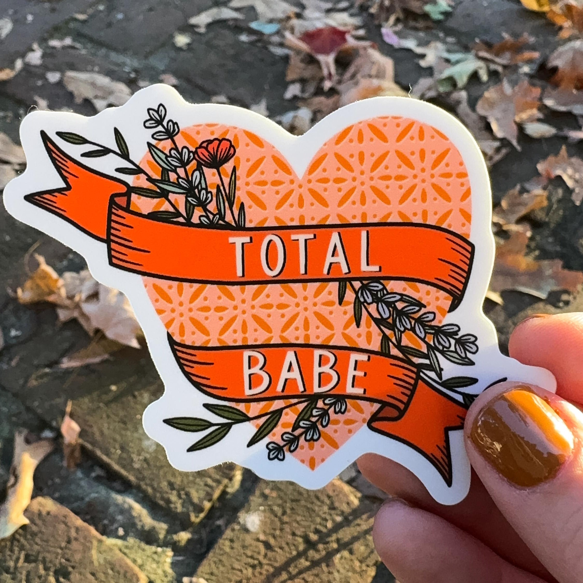 A hand holding a heart sticker with 'TOTAL BABE' written on a floral banner.