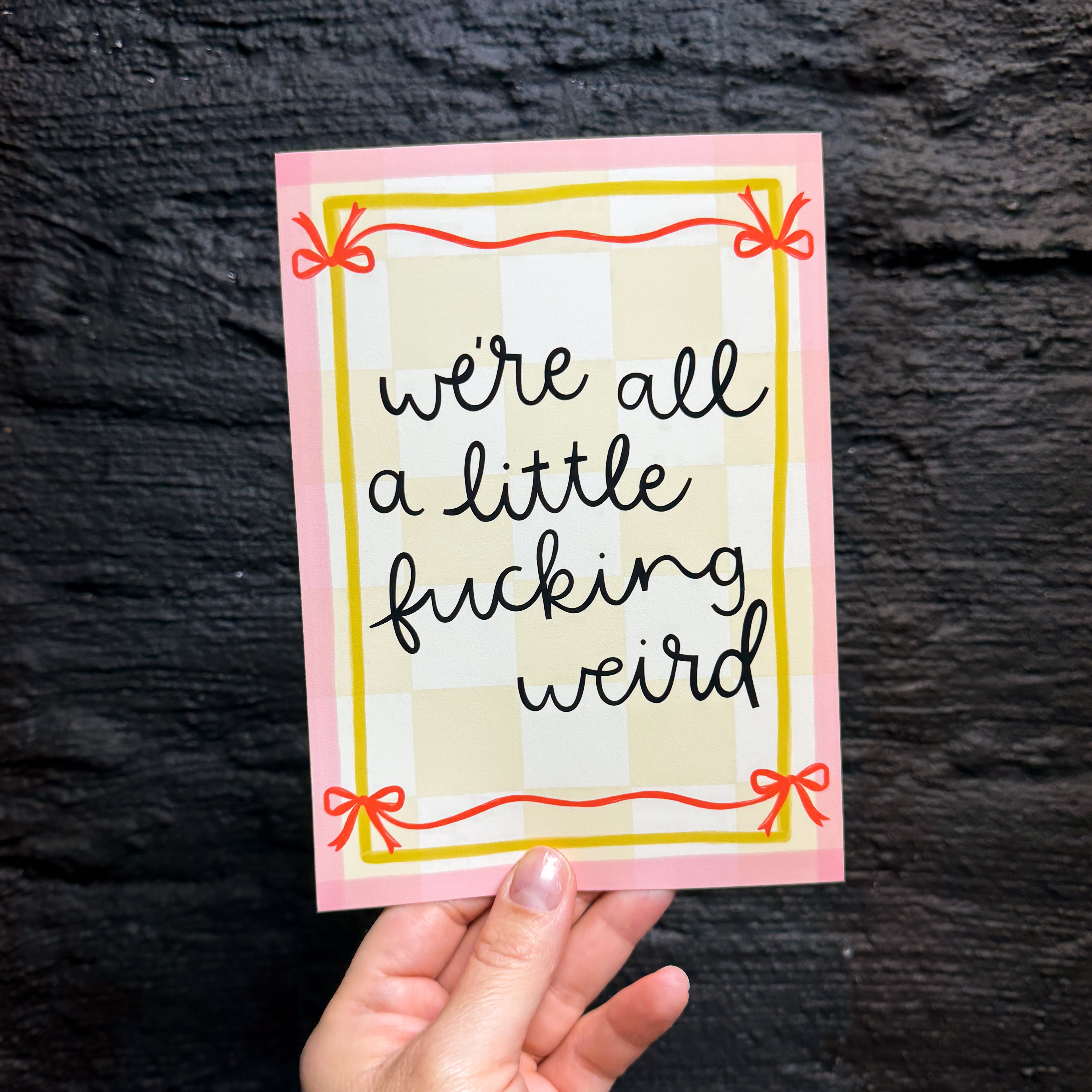 A hand holding a 5x7 art print that says 'we're all a little fucking weird' bordered by a yellow and pink frame with red bows. It has a pale yellow checkered background.