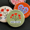 A round pinback button featuring a sage green background with red flowers and the text 'WHAT THE FUCK'. The pin is surrounded by other pin designs.