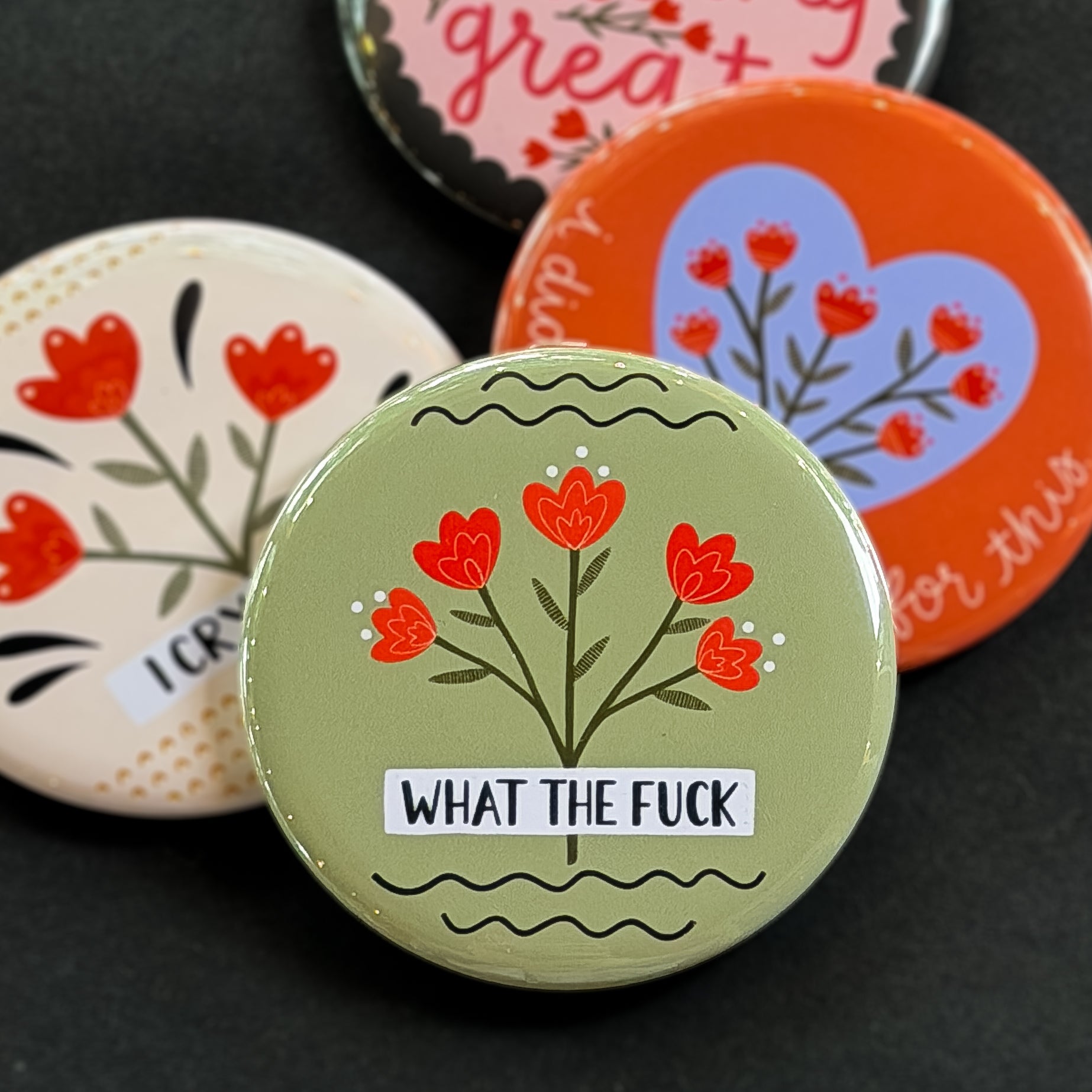 A round pinback button featuring a sage green background with red flowers and the text 'WHAT THE FUCK'. The pin is surrounded by other pin designs.