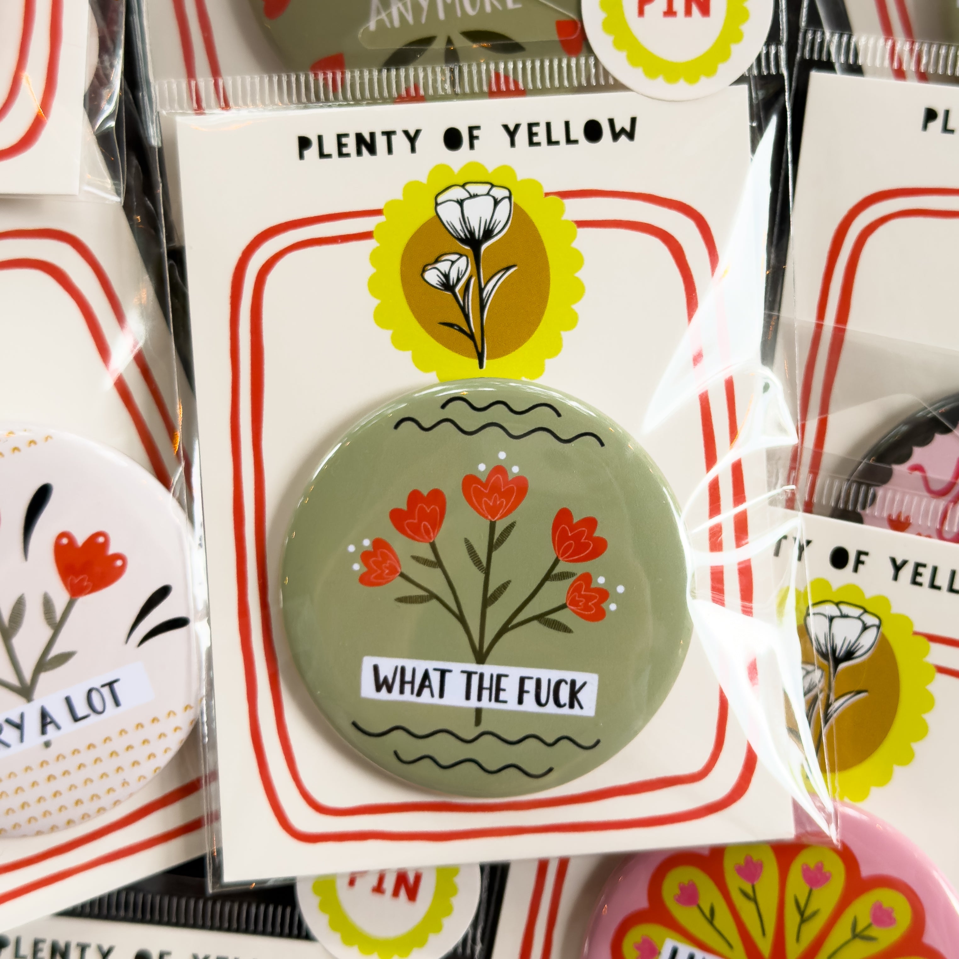 A round pinback button in clear plastic packaging, featuring a sage green background with red flowers and the text 'WHAT THE FUCK'. The pin is mounted on a white backing card with red borders and a yellow logo, under the brand name 'PLENTY OF YELLOW'.