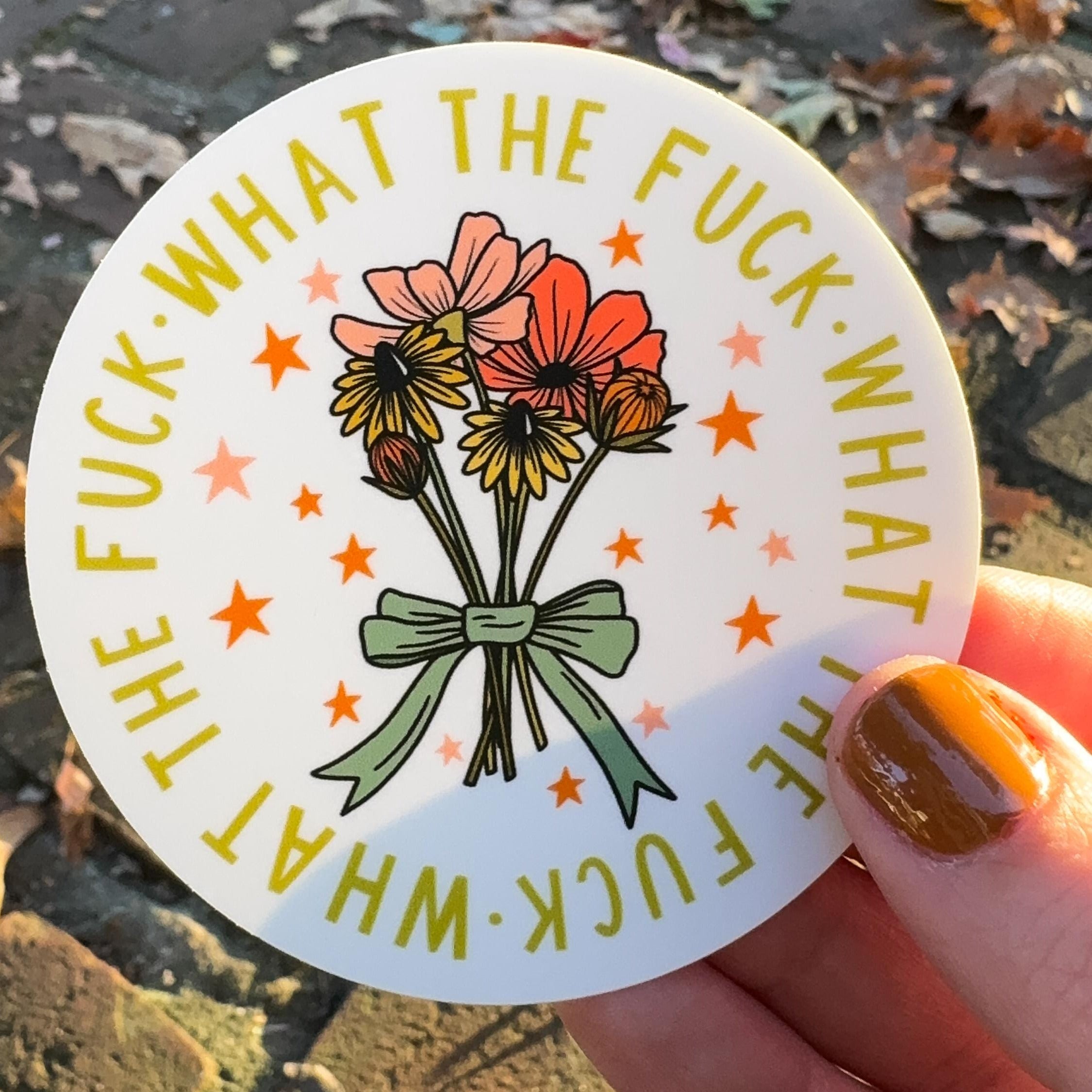 A hand holding a sticker with a floral design and text 'WHAT THE FUCK' on a white background