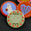 A round pinback button featuring a sage green background with red hearts around the edge and the text 'I DON'T KNOW WHAT THE FUCK IS HAPPENING ANYMORE'. The pin is surrounded by other pin designs.
