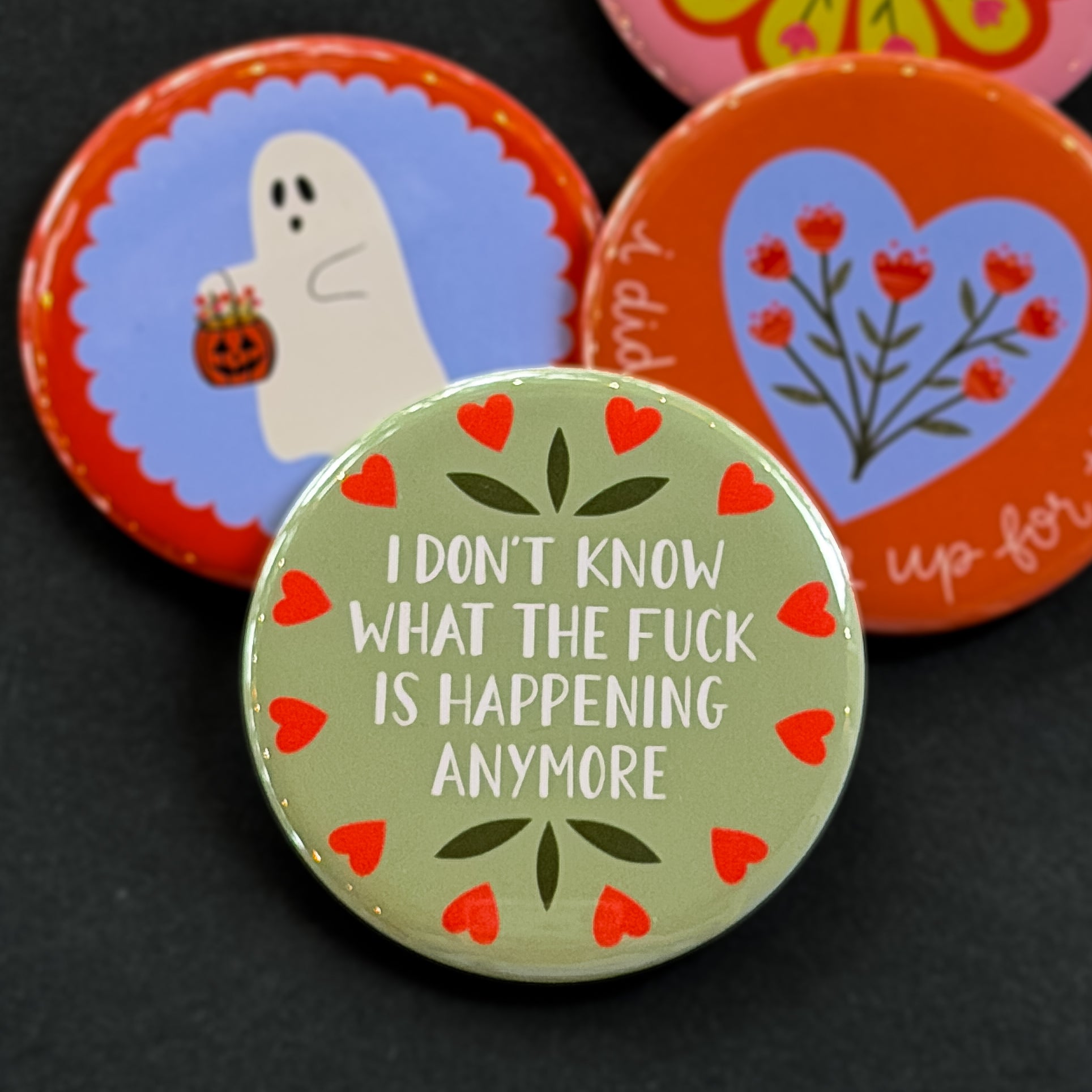 A round pinback button featuring a sage green background with red hearts around the edge and the text 'I DON'T KNOW WHAT THE FUCK IS HAPPENING ANYMORE'. The pin is surrounded by other pin designs.
