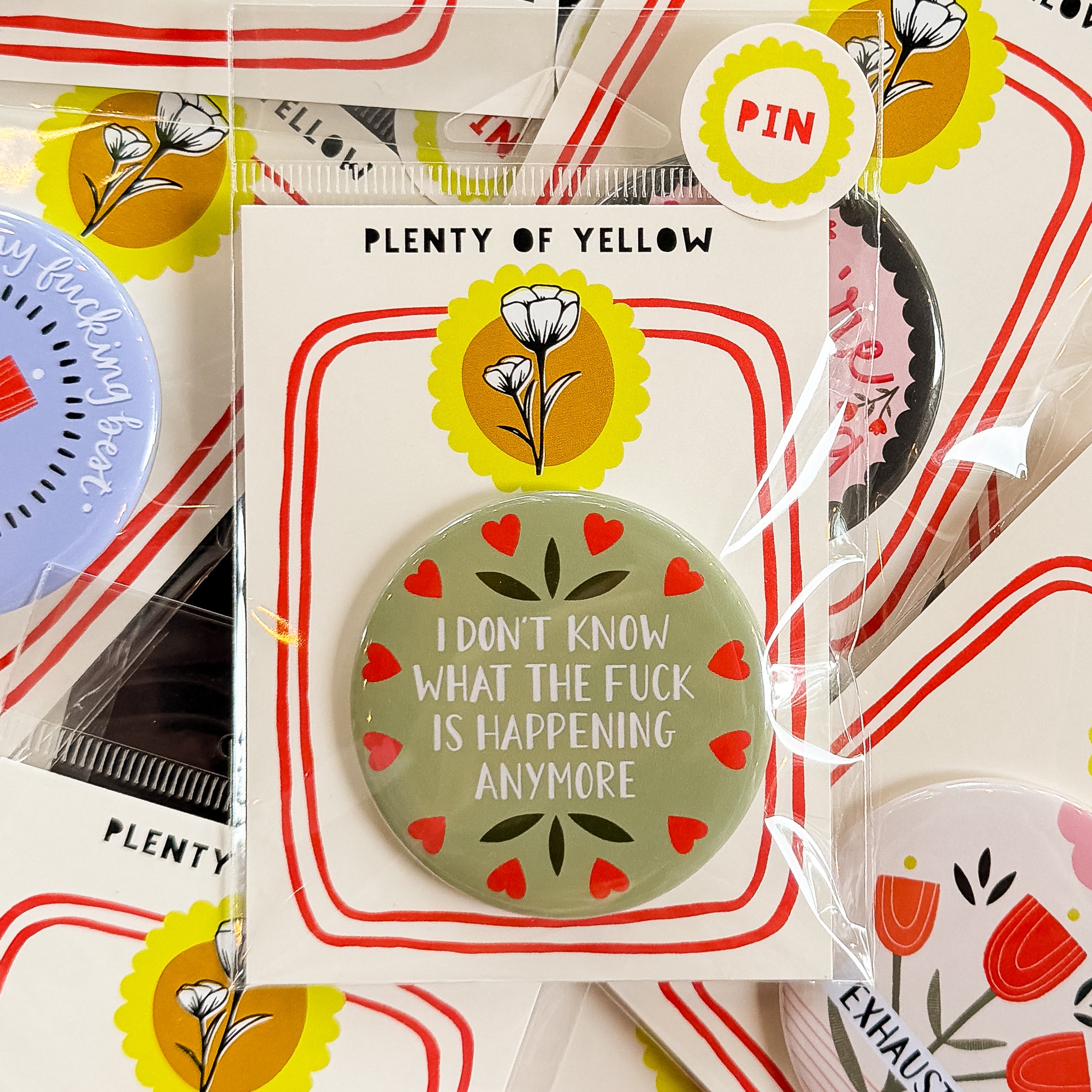 A round pinback button in clear plastic packaging, featuring a sage green background with red hearts around the edge and the text 'I DON'T KNOW WHAT THE FUCK IS HAPPENING ANYMORE'. The pin is mounted on a white backing card with red borders and a yellow logo, under the brand name 'PLENTY OF YELLOW'.
